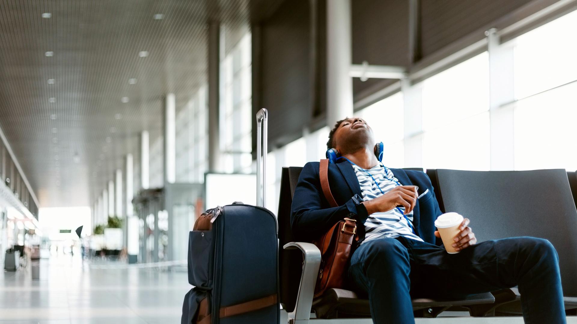 Ein junger Mann sitzt müde, mit geschlossenen Augen, Kopfhörern und einem Kaffebecher neben seinem Gepäck am Flughafen und wartet.