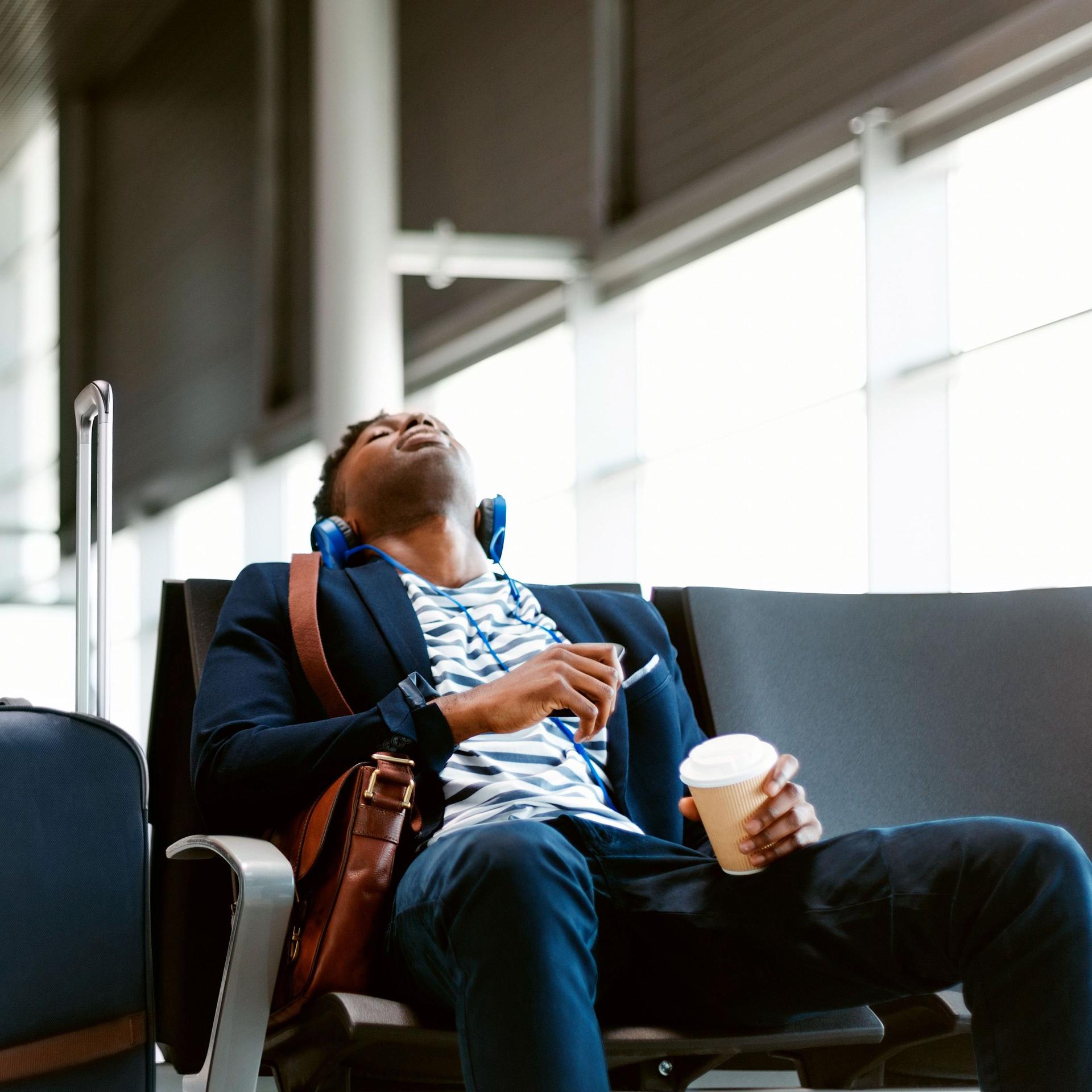 Ein junger Mann sitzt müde, mit geschlossenen Augen, Kopfhörern und einem Kaffebecher neben seinem Gepäck am Flughafen und wartet.