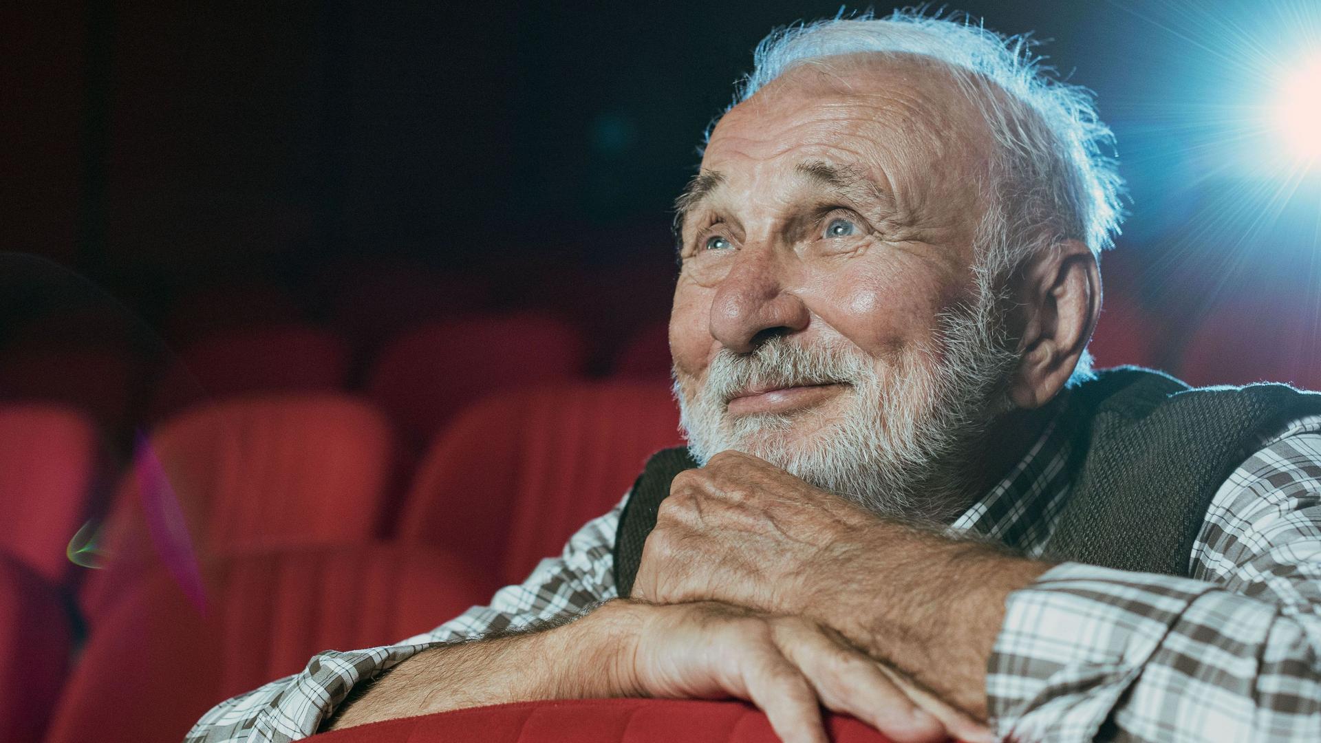 Ein Senior mit grauen Haaren und Bart sitzt in einem Kino und schaut lächelnd einen Film.