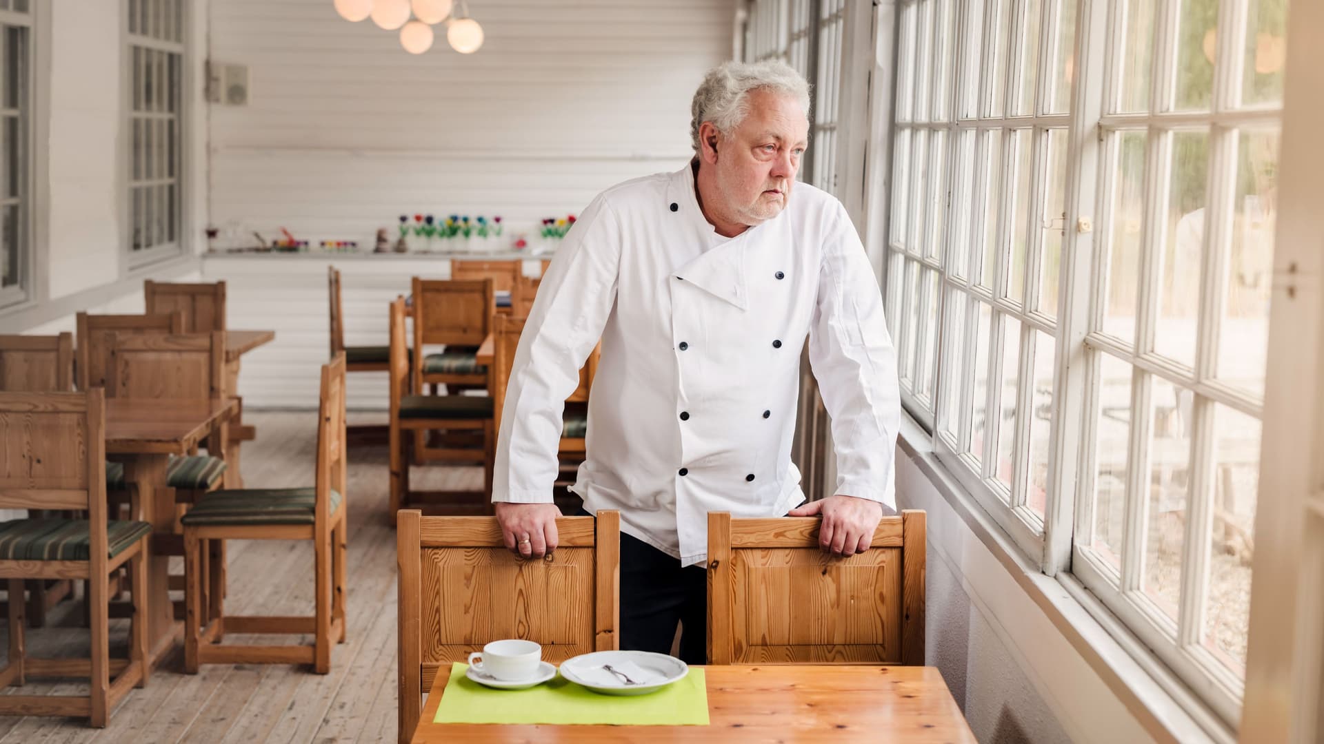 Ein Mann in weißer Kochjacke steht in einem leerem Restaurant an einem Tisch. Er blickt traurig aus dem Fenster.