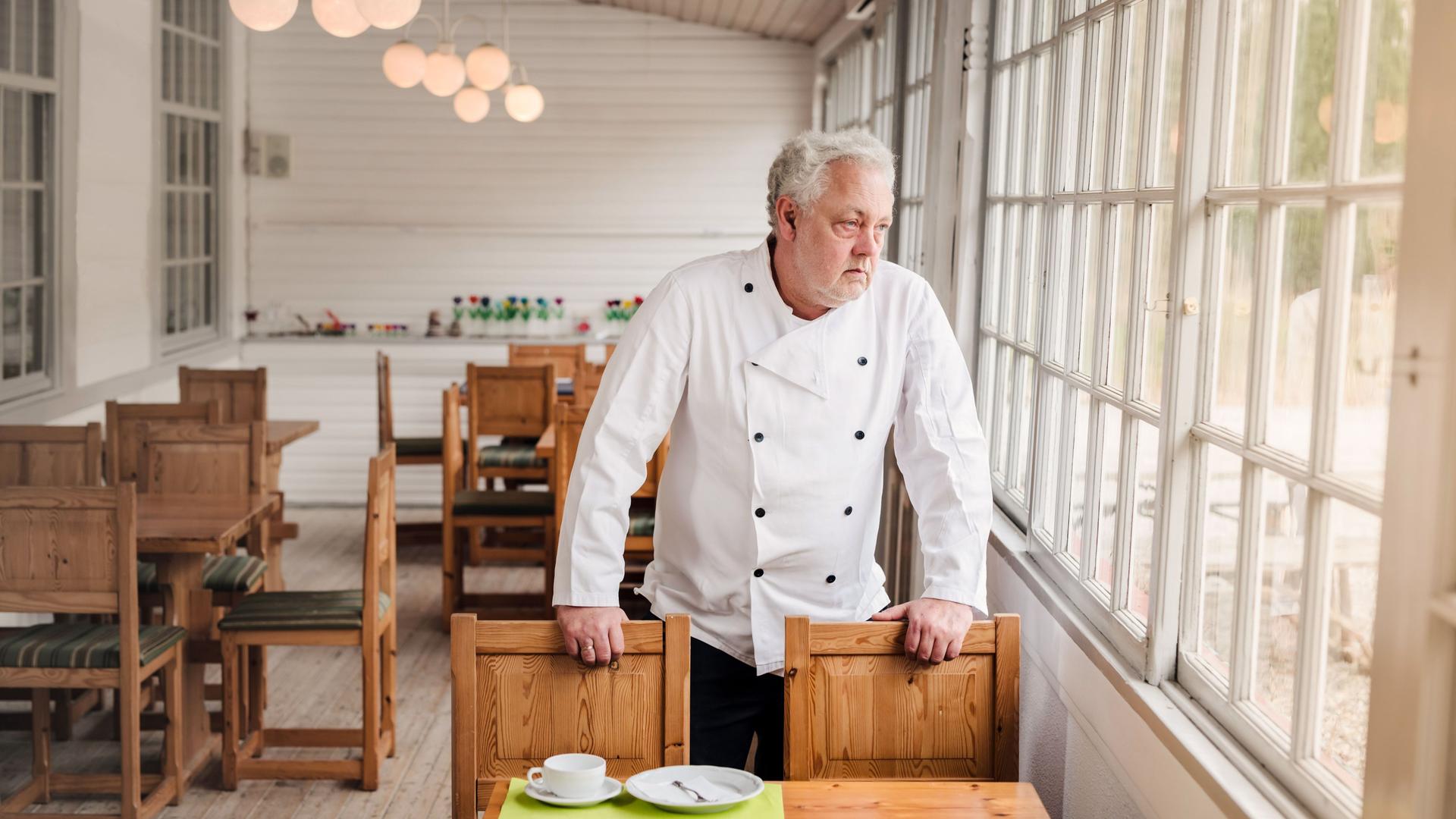 Ein Mann in weißer Kochjacke steht in einem leerem Restaurant an einem Tisch. Er blickt traurig aus dem Fenster.