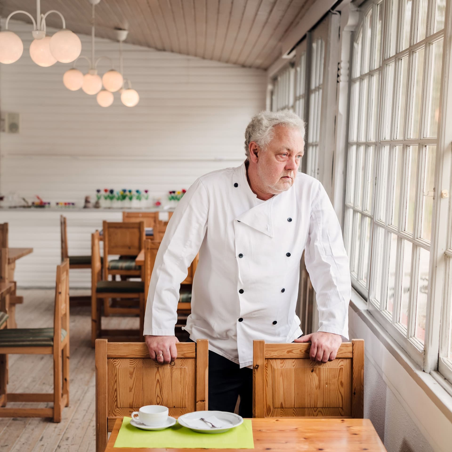 Ein Mann in weißer Kochjacke steht in einem leerem Restaurant an einem Tisch. Er blickt traurig aus dem Fenster.