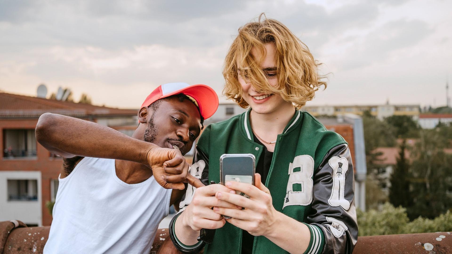 Zwei junge, erwachsene Männer, in sportlich modischer, Freizeitkleidung, stehen auf dem Dach in einer Wohnsiedlung und sehen zusammen auf den Screen eines Smartphones. 