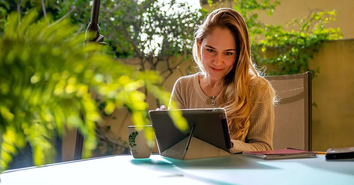 Eine Frau sitzt auf einer Terrasse in einem grün bewachsenen Garten und schaut lächelnd auf ihr Tablet, welches auf dem Tisch steht. 