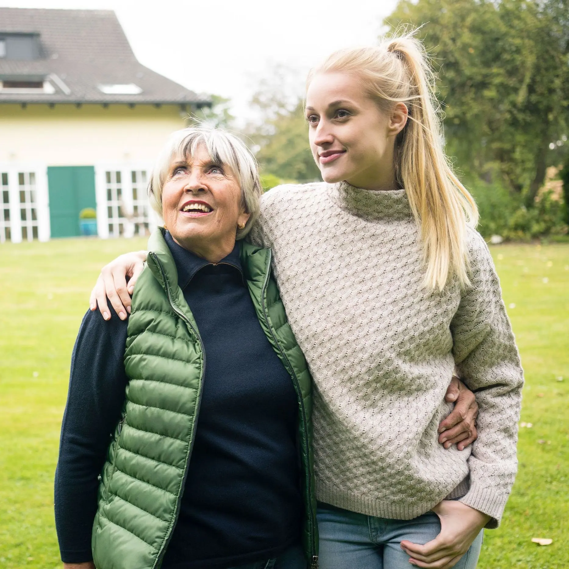 Eine junge Frau mit langem blonden Pferdeschwanz legt ihren Arm um ihre Großmutter. Sie stehen auf der Wiese eines Grundstücks. Im Hintergrund steht ein schönes altes Wohnhaus. Beide lächeln.