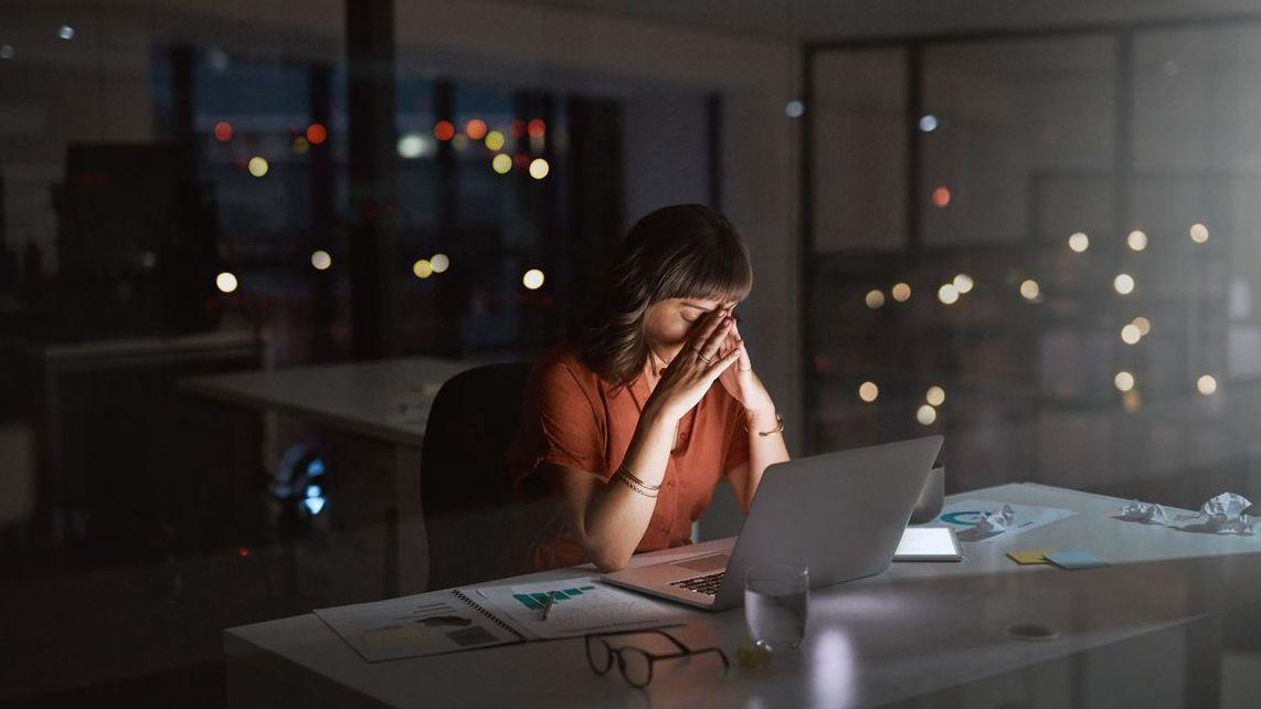 Eine Frau sitzt spät abends in einem dunklen Büro vor einem Laptop . Sie stützt ihren Kopf müde auf die Hände.