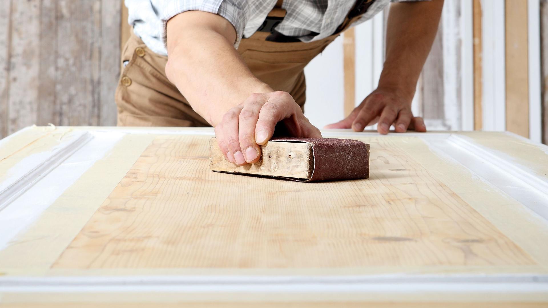Schreiner mit Overall und Mütze, der in einer Schreinerei Holz bearbeitet und eine Holztür mit Schleifpapier abschleift.