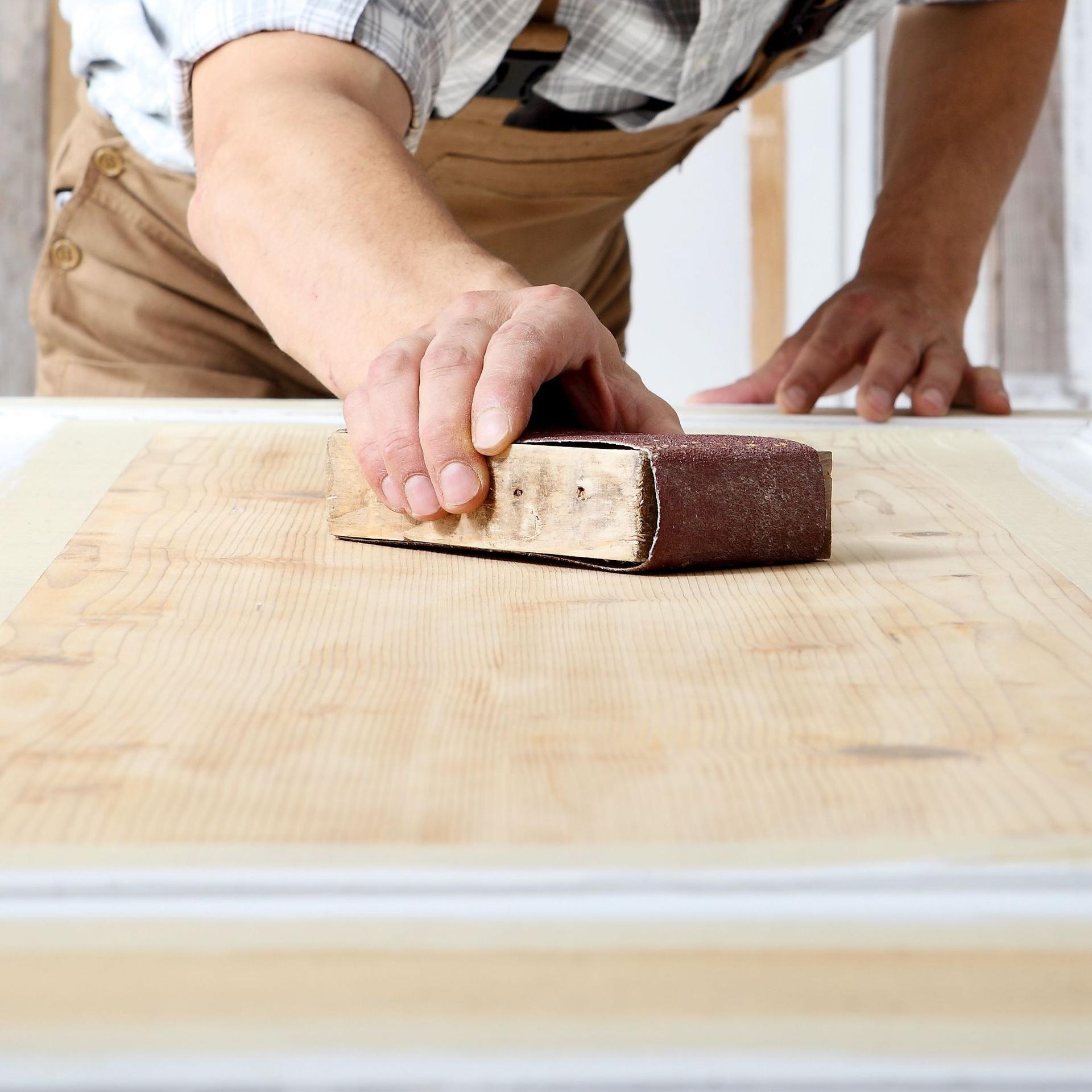 Schreiner mit Overall und Mütze, der in einer Schreinerei Holz bearbeitet und eine Holztür mit Schleifpapier abschleift.