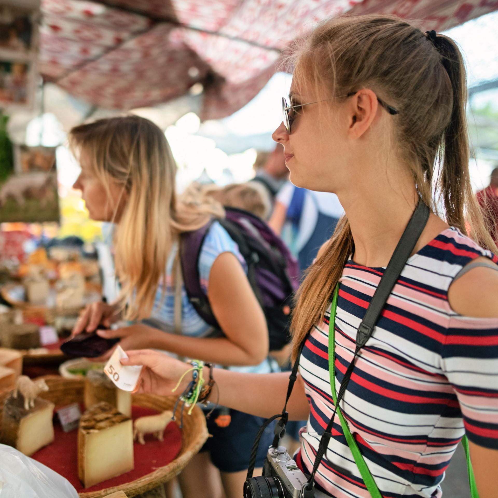 Eine junge Frau mit Sonnenbrille und Kamera um den Hals bezahlt an einem Marktstand.