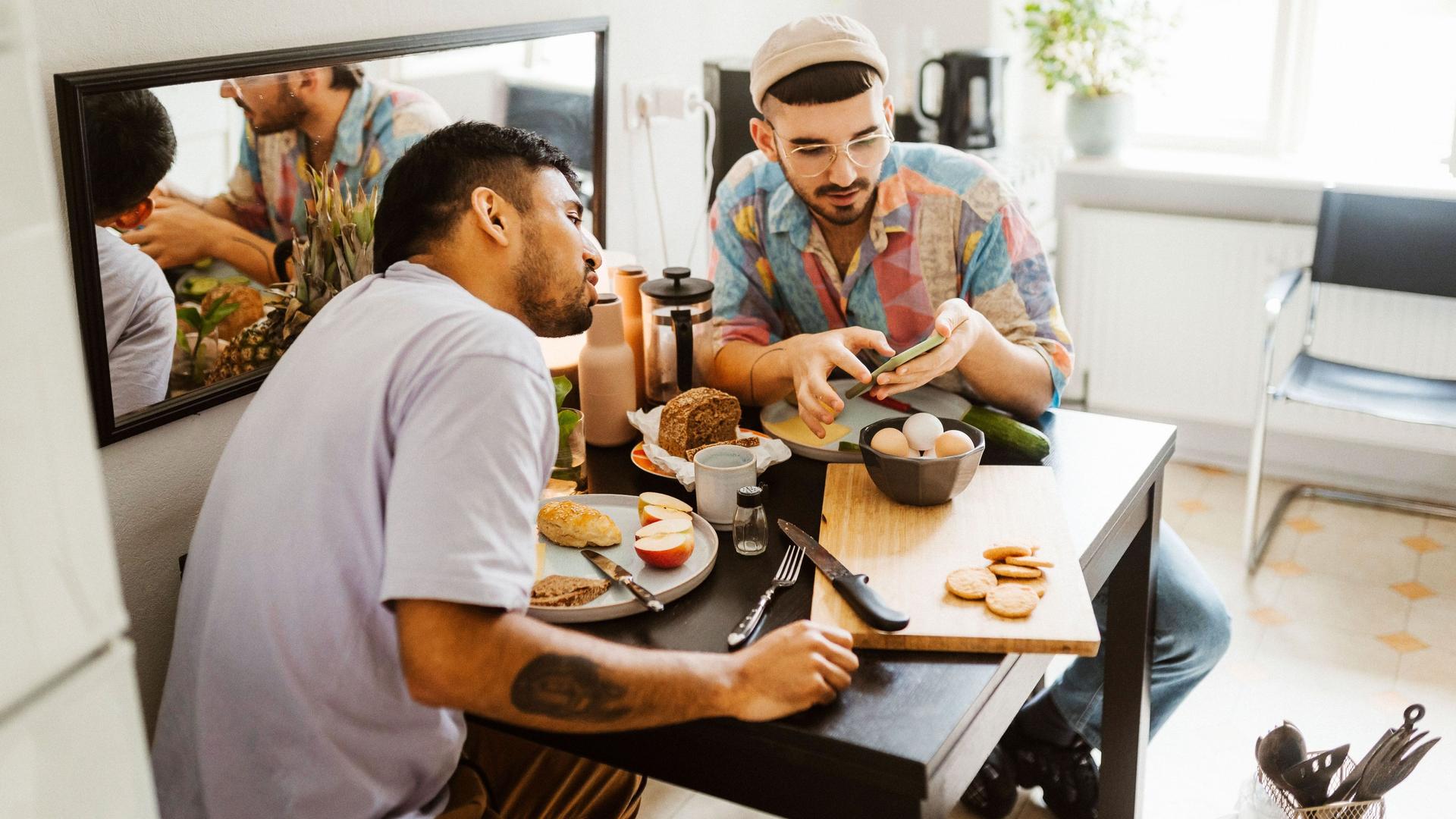 Ein schwules Paar sitzt in der Küche am Frühstückstisch. Der eine zeigt dem anderen etwas auf seinem Smartphone.