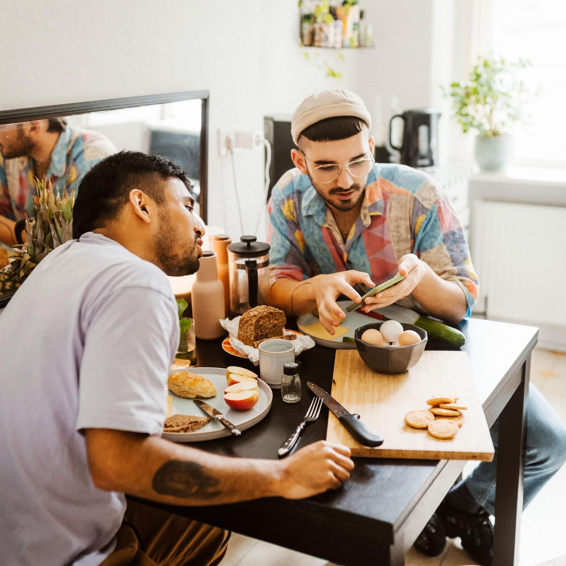 Ein schwules Paar sitzt in der Küche am Frühstückstisch. Der eine zeigt dem anderen etwas auf seinem Smartphone.