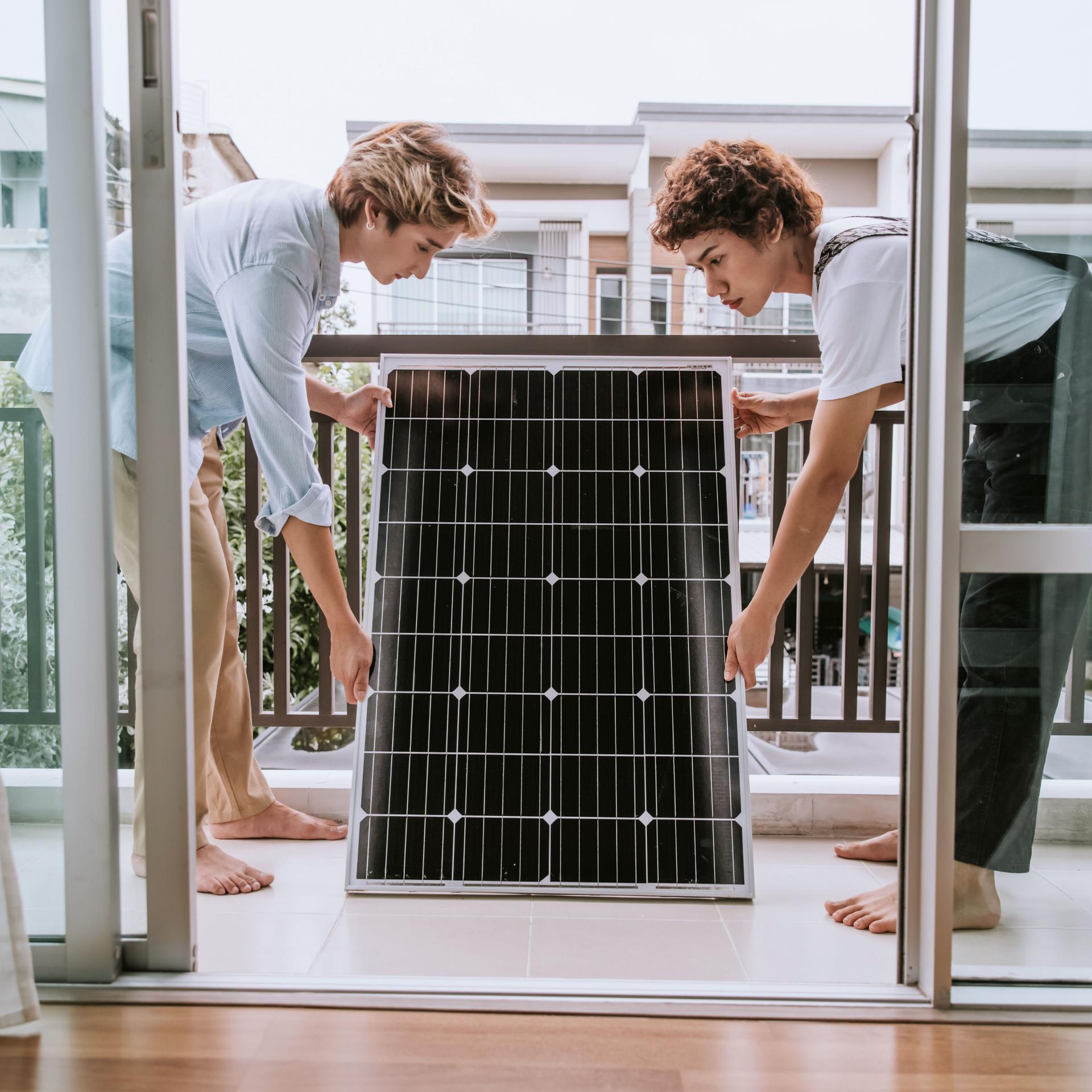 Zwei Personen stehen mit einem Solarpanel in den Händen auf dem Balkon eines Hauses.
