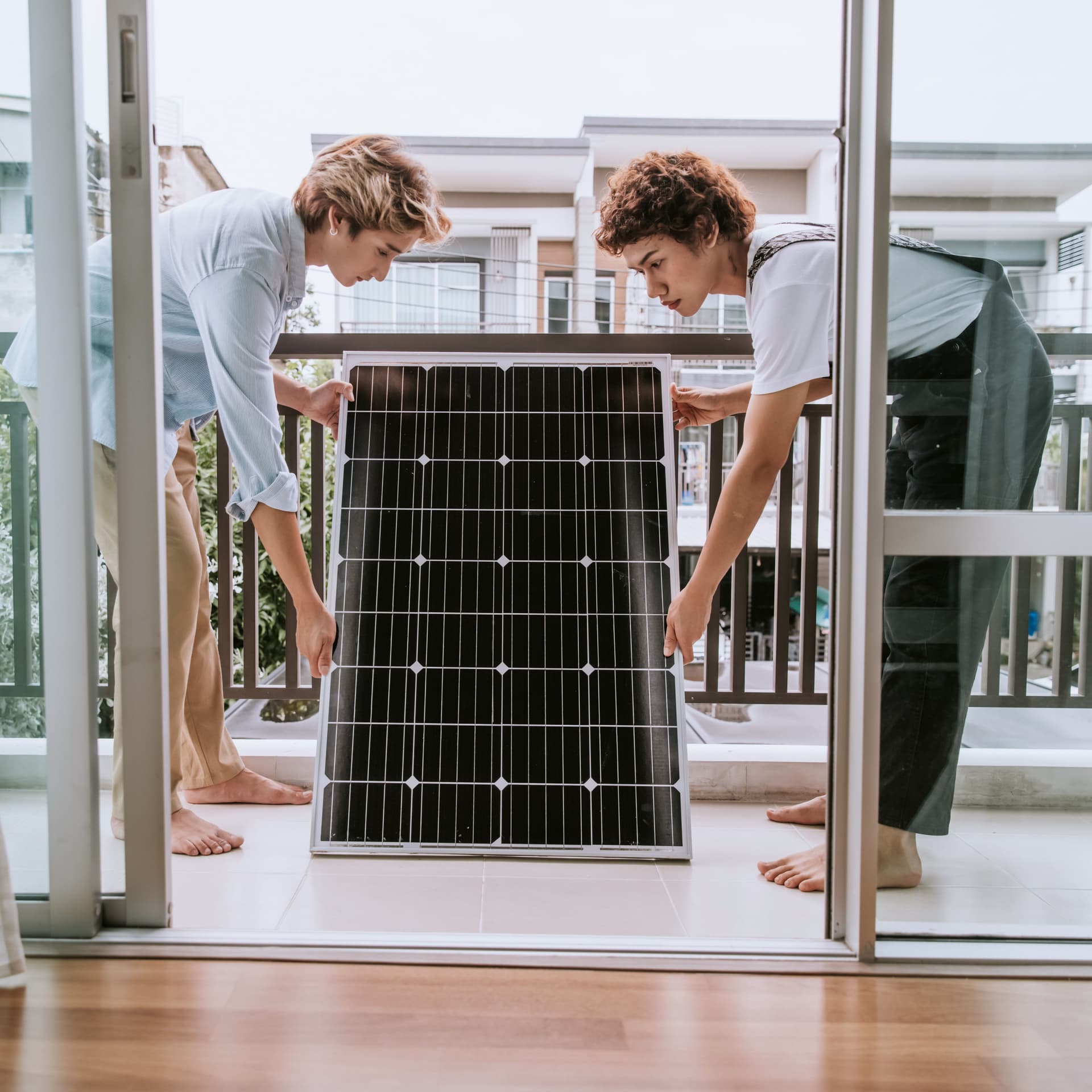 Zwei Personen stehen mit einem Solarpanel in den Händen auf dem Balkon eines Hauses.