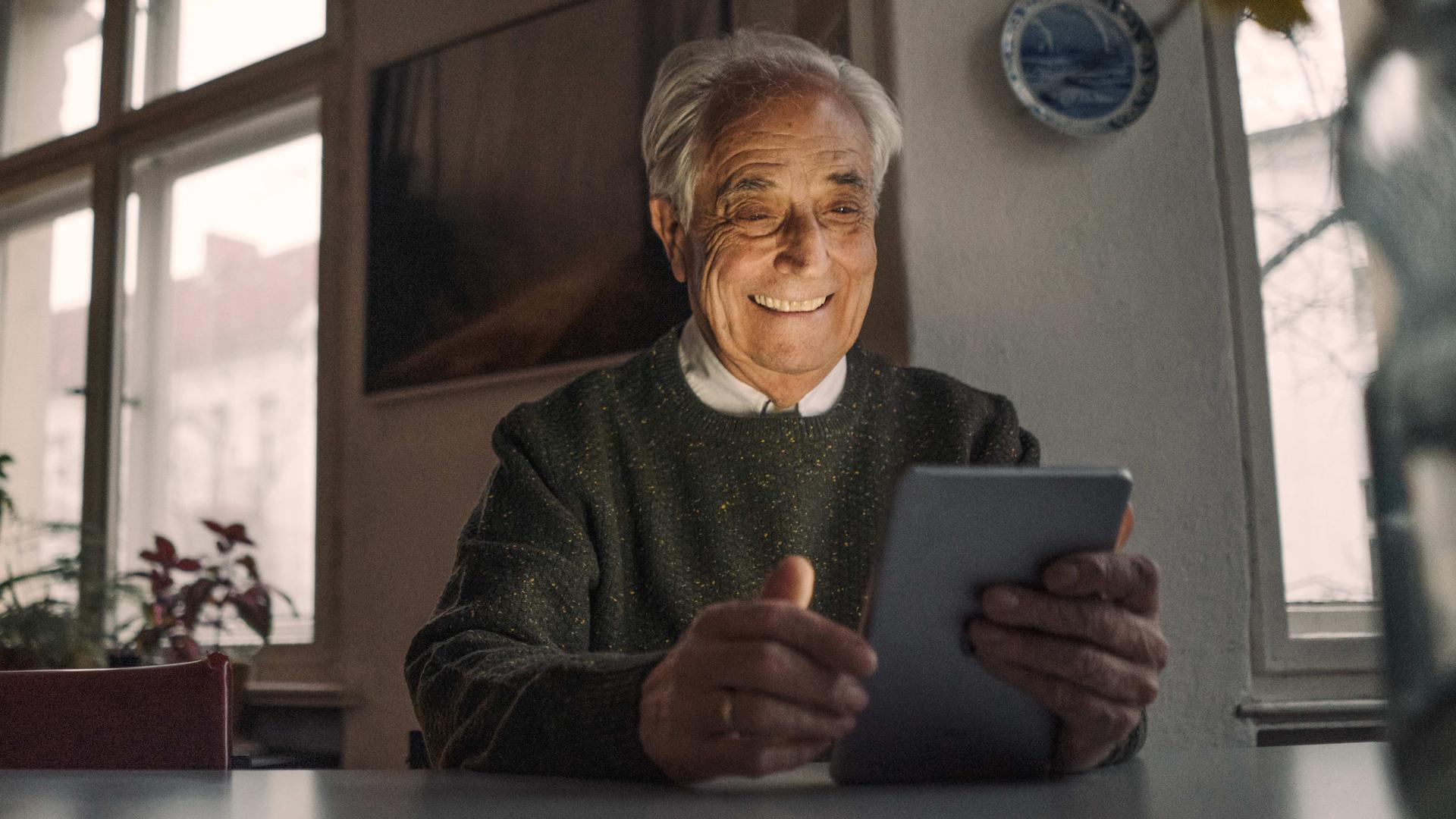 Älterer Mann mit weißen Haaren sitzt am Tisch in einer abendlichen Wohnung und hält lächelnd ein Tablet