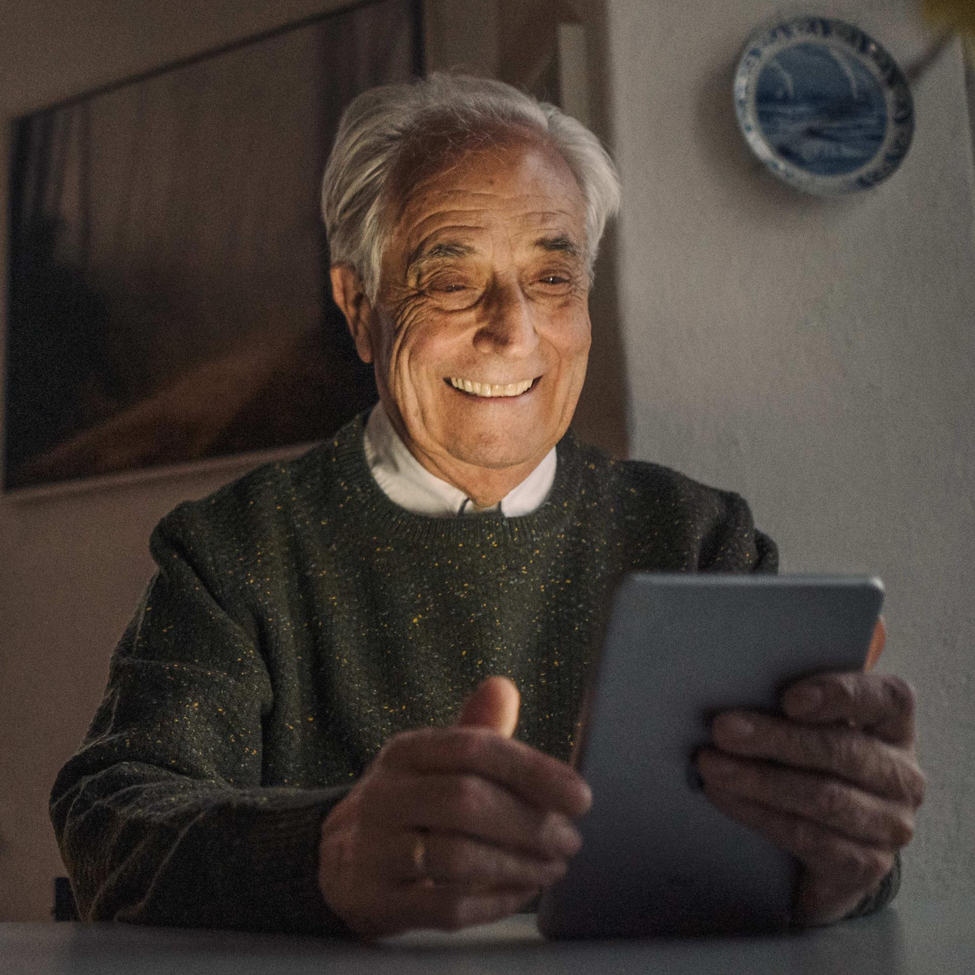 Älterer Mann mit weißen Haaren sitzt am Tisch in einer abendlichen Wohnung und hält lächelnd ein Tablet