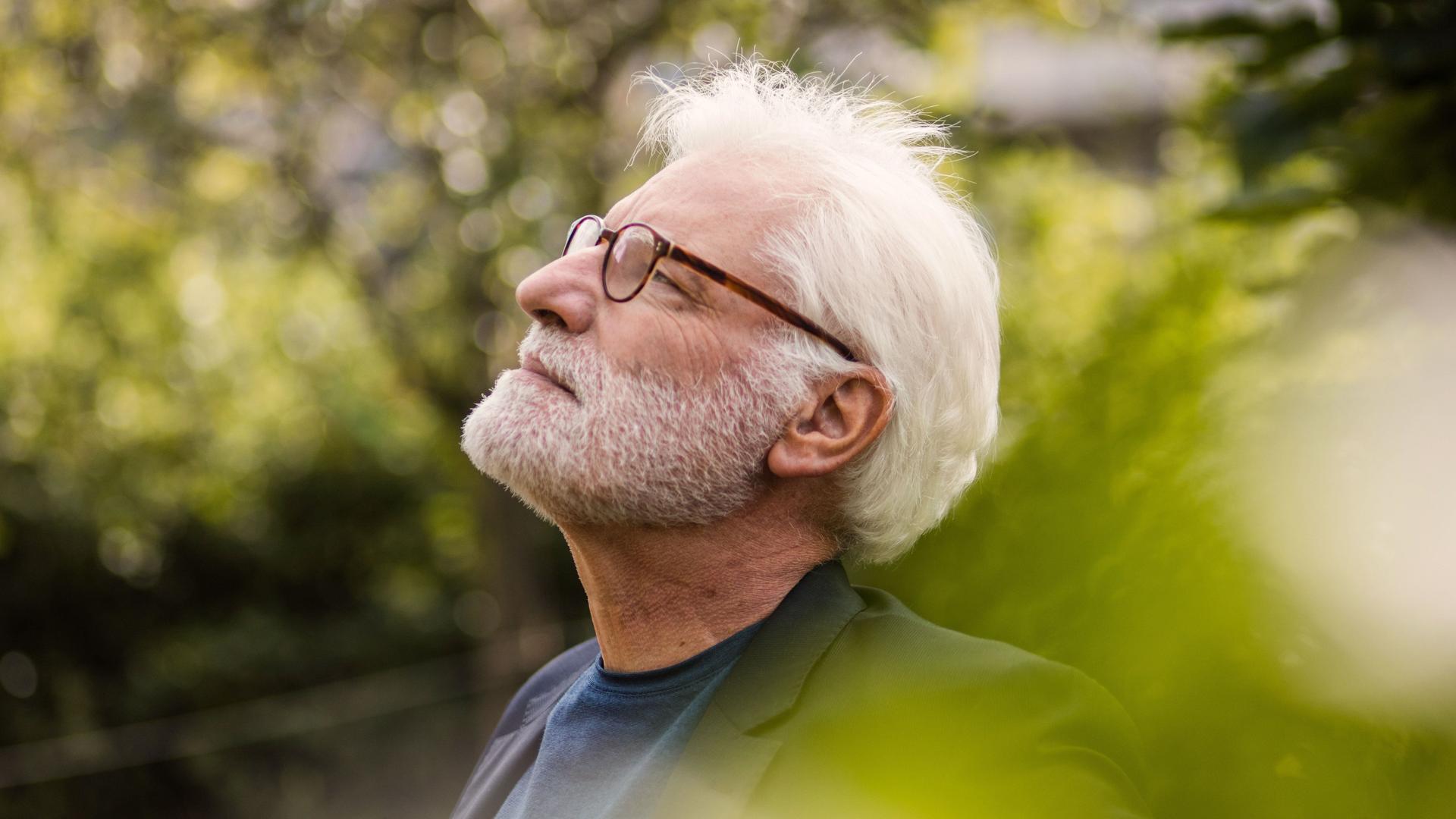 Bärtiger älterer Mann mit weißem Haar steht nachdenklich im Garten und schaut in den Himmel.