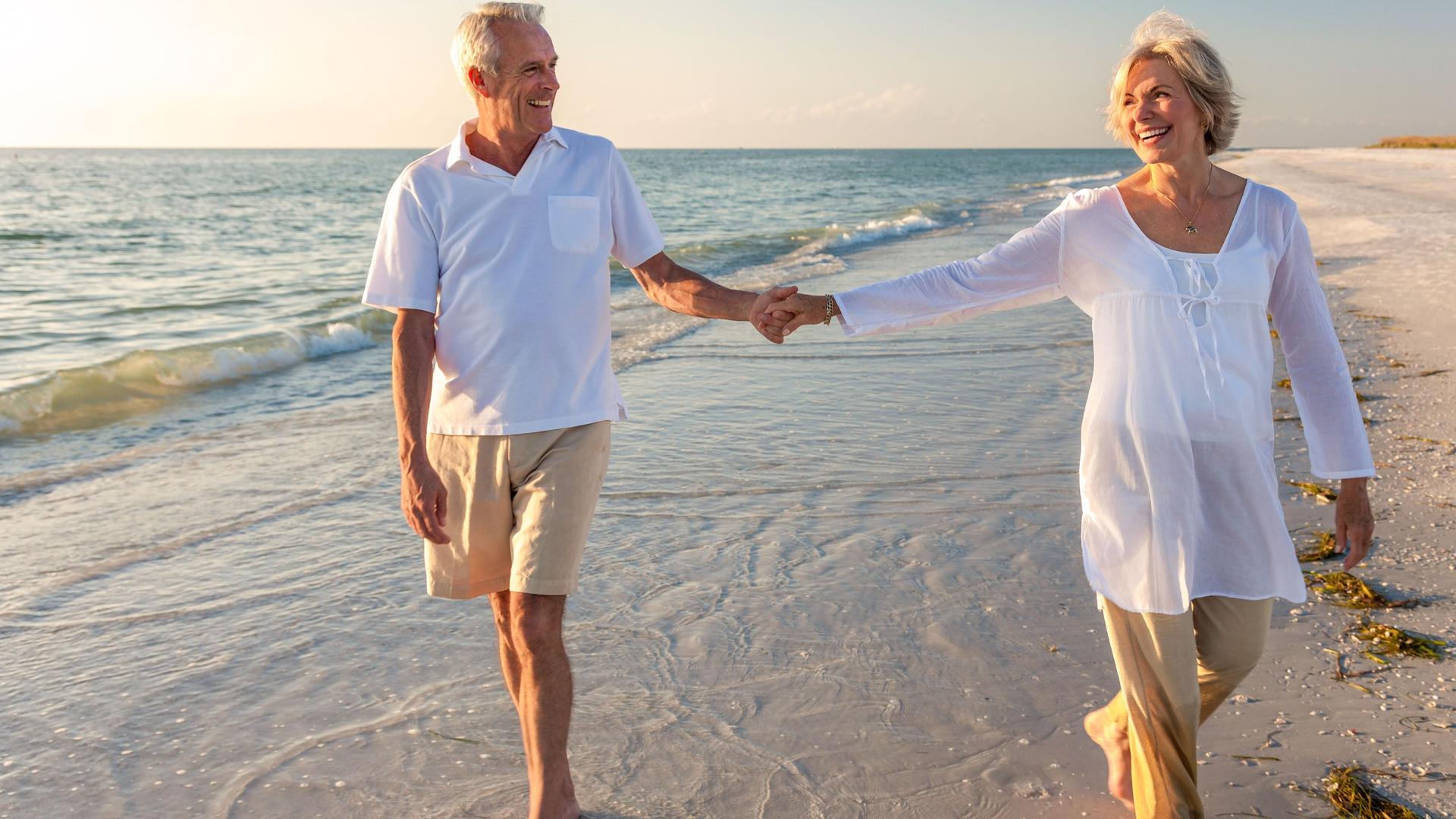Lachendes Seniorenpaar läuft Hand in Hand durch das flache Meerwasser am Strand.