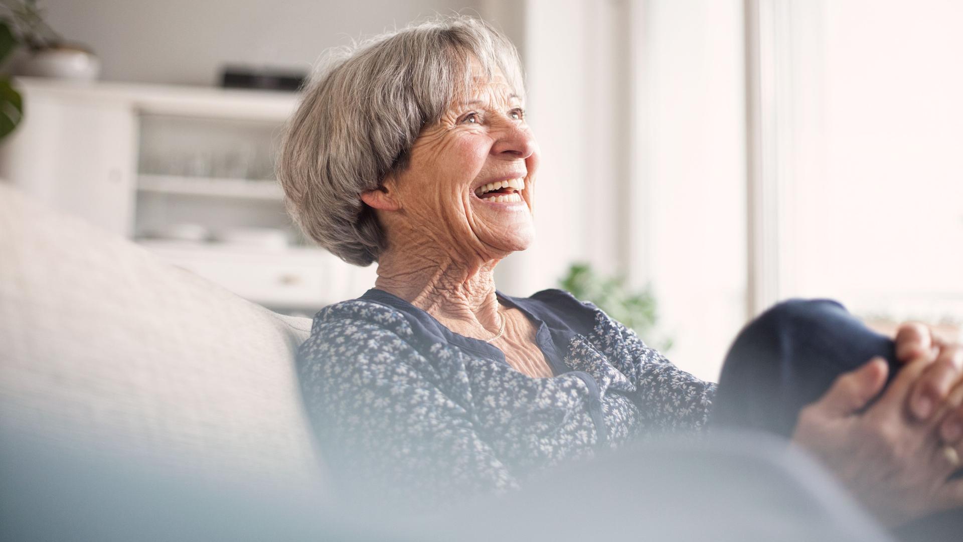 Porträt einer lachenden älteren Frau, die auf einem grauen Sofa sitzt.