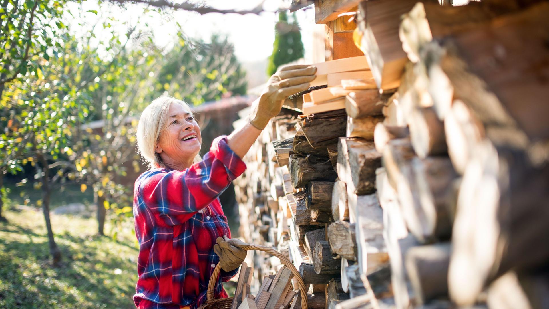 Seniorin lädt draußen bei Sonnenschein lachend Holzscheite von einem Holzstapel in einen Korb