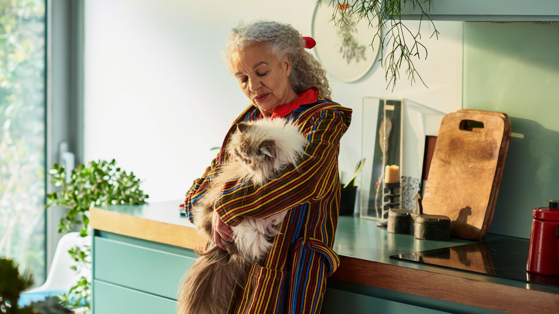 Seniorin lehnt an einer Küchenzeile und hält eine Katze auf dem Arm.