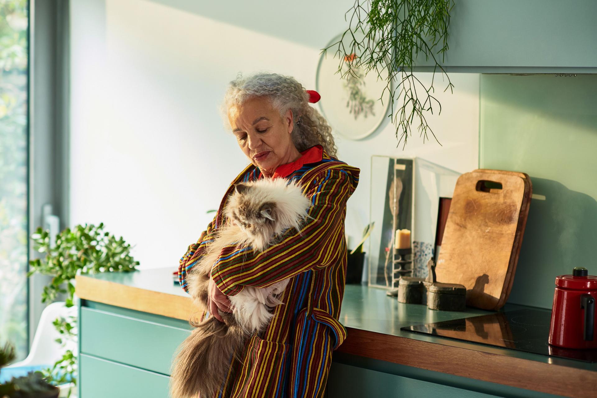 Seniorin lehnt an einer Küchenzeile und hält eine Katze auf dem Arm.