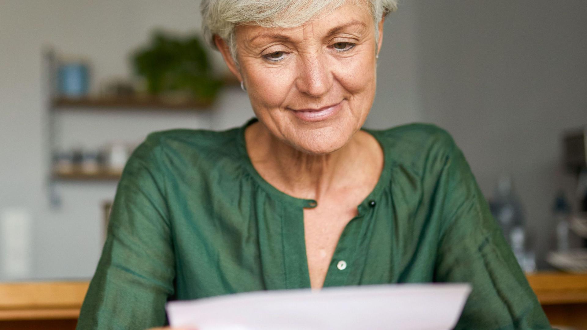 Eine Seniorin sitzt in einer Wohnung an einem Tisch und liest lächelnd einen Brief. Neben ihr steht eine Tasse.
