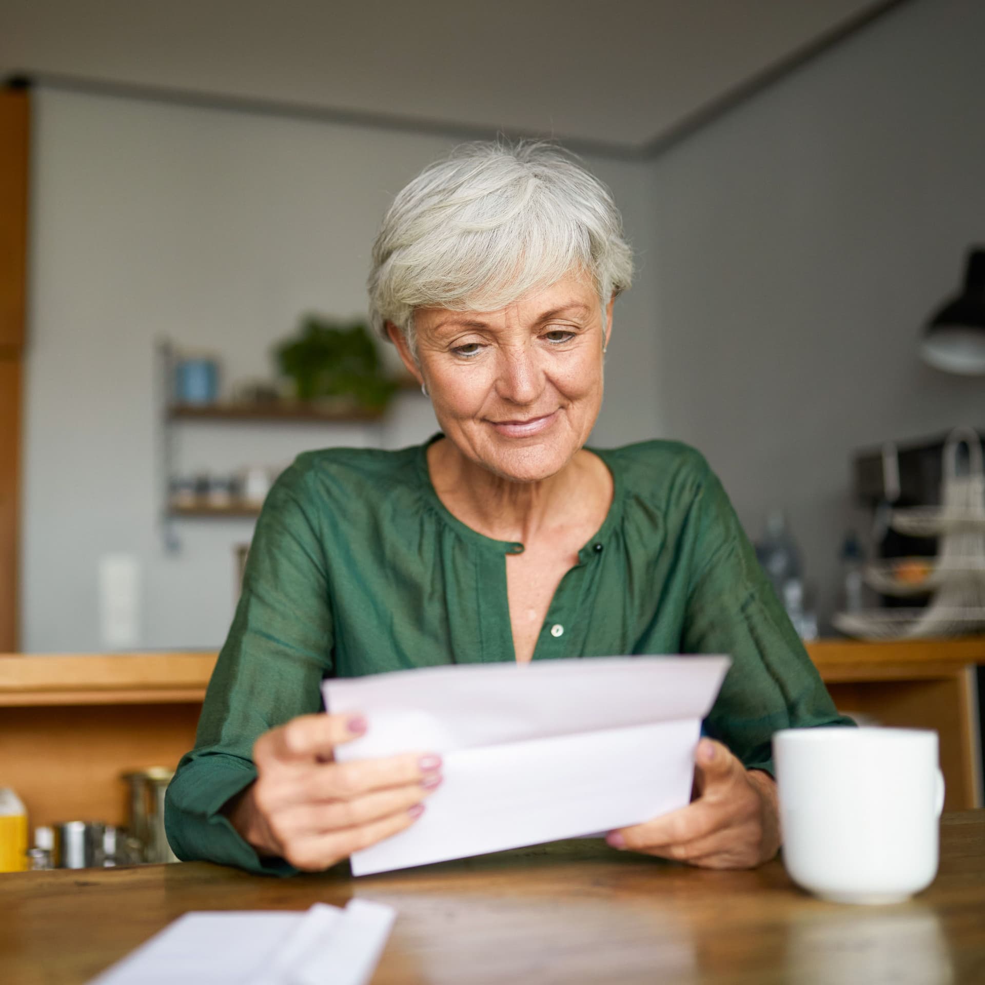 Eine Seniorin sitzt in einer Wohnung an einem Tisch und liest lächelnd einen Brief. Neben ihr steht eine Tasse.