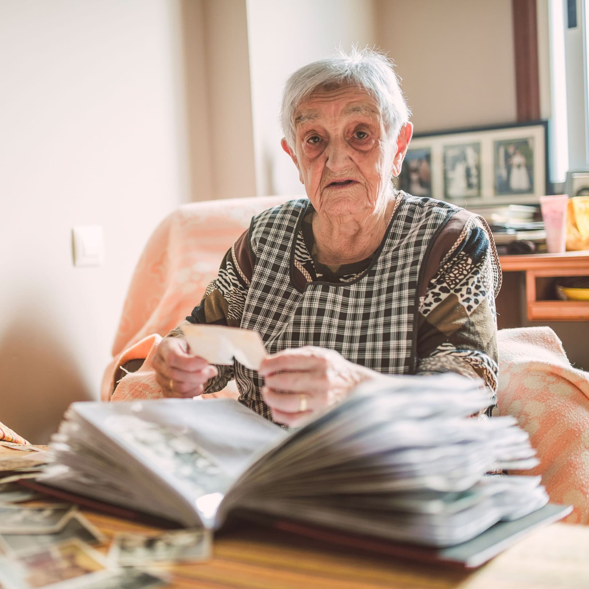 Eine Seniorin sitzt mit einem Fotoalbum vor einem Bett und blickt in die Kamera