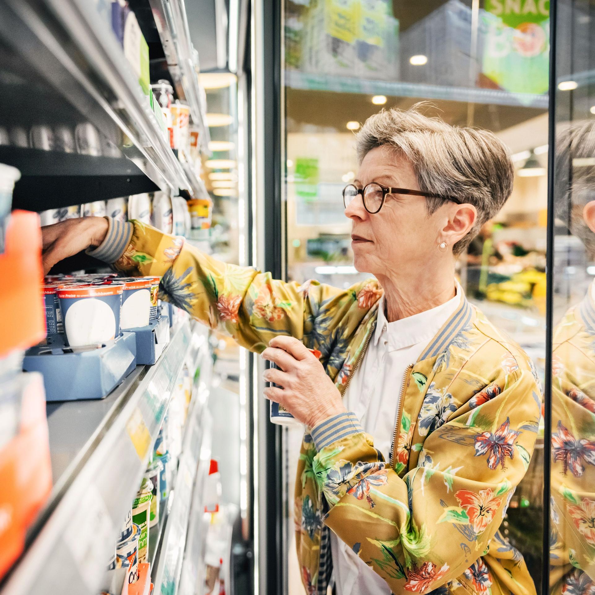Eine ältere Frau im Supermarkt, die Lebensmittel aus einem Kühlschrank herausholt.