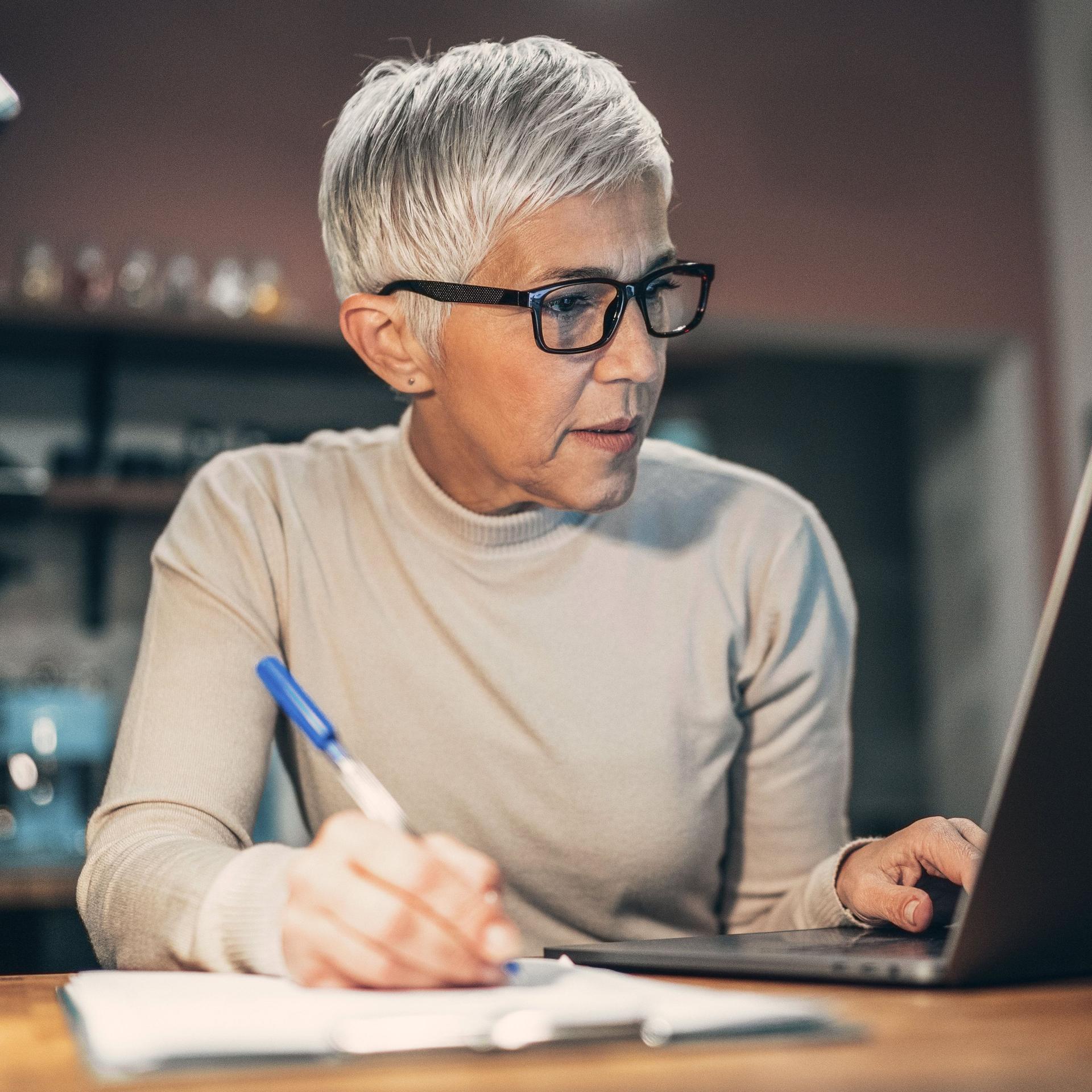 Ältere Frau sitzt vor einem Laptop in der Küche. Sie hält einen Stift in der rechten Hand, Papier liegt auf dem Tisch.