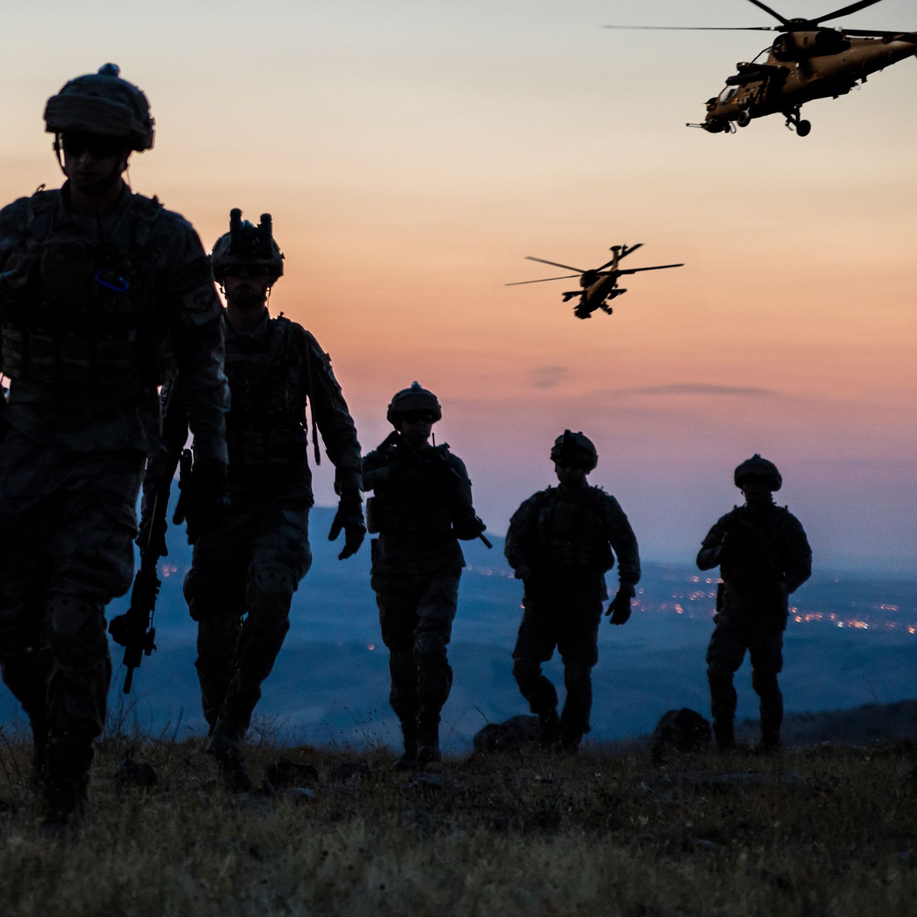 Silhouetten von fünf Soldaten in voller Ausrüstung, die einen Hügel hinaufgehen, während zwei Hubschrauber am Dämmerungshimmel fliegen und eine Stadt in der Ferne leuchtet.