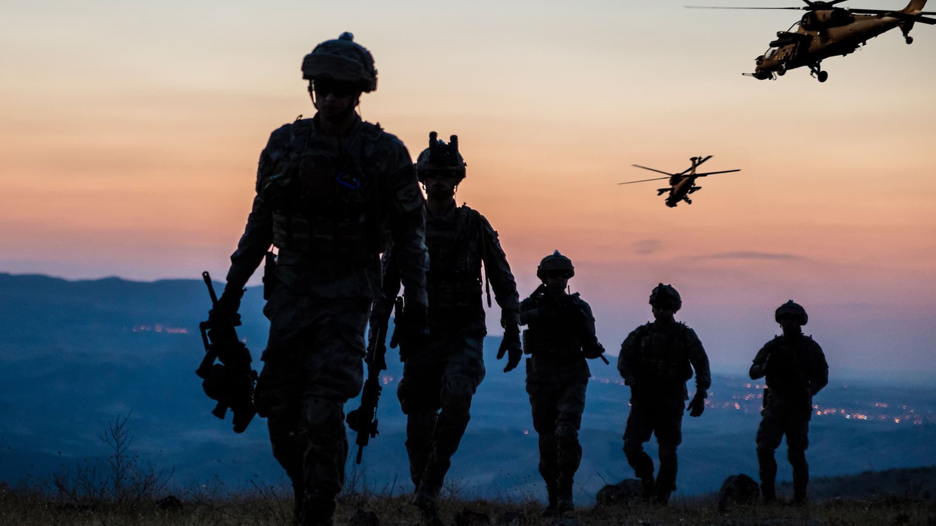 Silhouetten von fünf Soldaten in voller Ausrüstung, die einen Hügel hinaufgehen, während zwei Hubschrauber am Dämmerungshimmel fliegen und eine Stadt in der Ferne leuchtet.