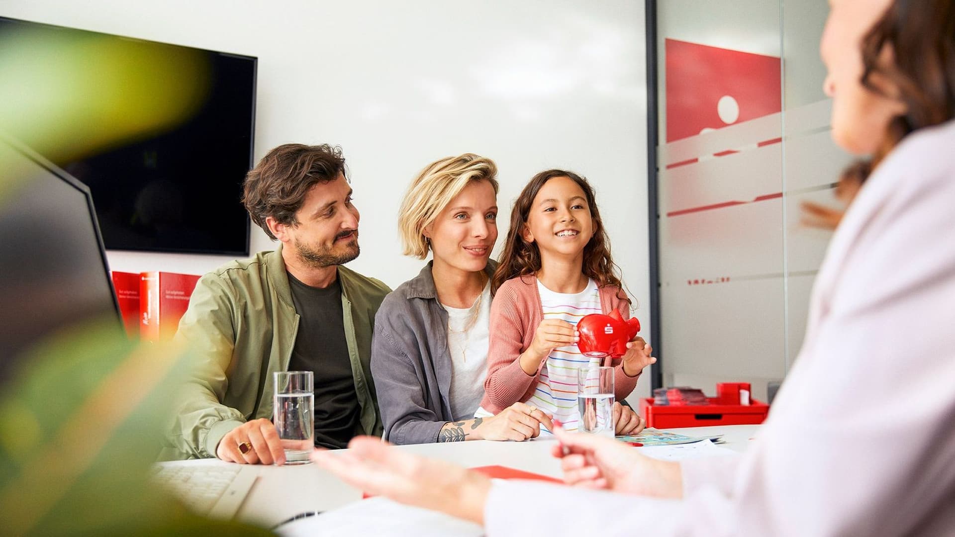 Eine dreiköpfige Familie sitzen an einem Tisch in einer Sparkassenfiliale. Das Kind hält ein Sparschwein in den Händen. Am anderen Ende des Tisches sieht man in Seitenansicht eine Beraterin.