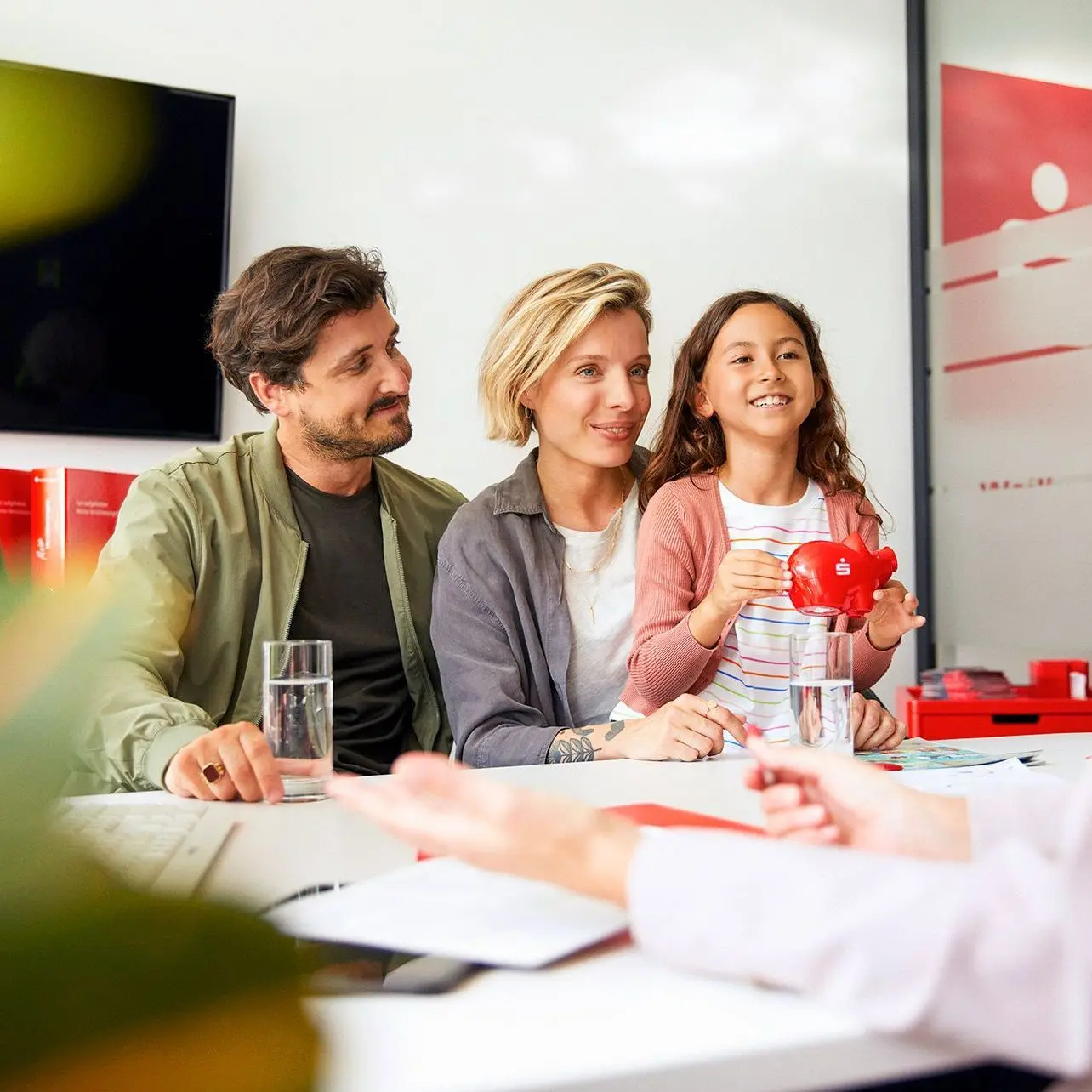 Eine dreiköpfige Familie sitzen an einem Tisch in einer Sparkassenfiliale. Das Kind hält ein Sparschwein in den Händen. Am anderen Ende des Tisches sieht man in Seitenansicht eine Beraterin.