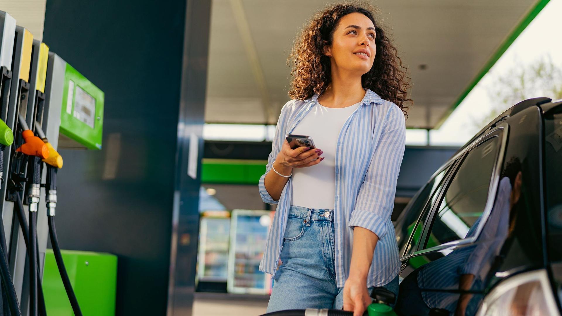 Eine Frau mit lockigen Haaren und sommerlicher Kleidung tankt an einer Tankstellenzapfsäule Benzin. Sie hält ein Smartphone in der Hand und schaut nach oben auf die Preistafel. 