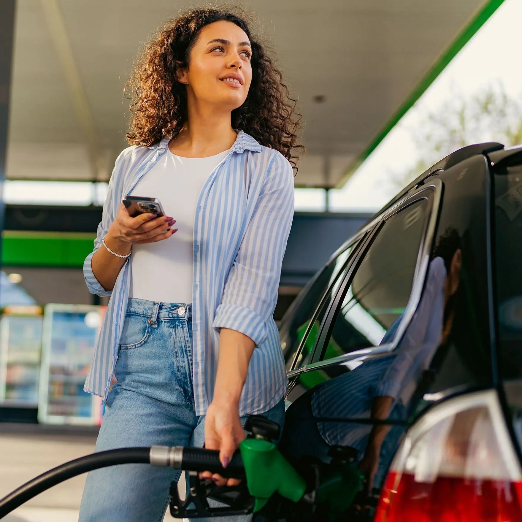 Eine Frau tankt an einer Tankstellenzapfsäule Benzin. Sie hält ein Smartphone in der Hand und schaut nach oben auf die Preistafel. 