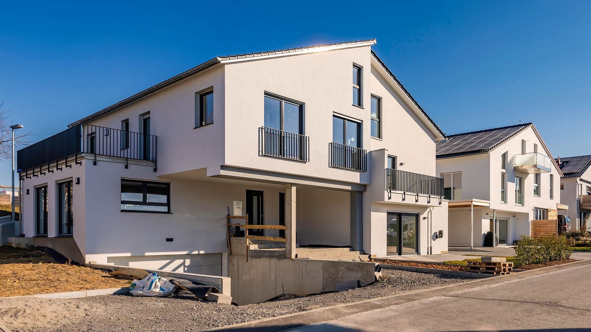 Eine Außenaufnahme eines Straßenzuges mit neugebauten Einfamilienhäusern. Ein weißes Haus ist zentral im Bild. Ähnliche Häuser reihen sich anhand einer Fluchtlinie hintereinander.