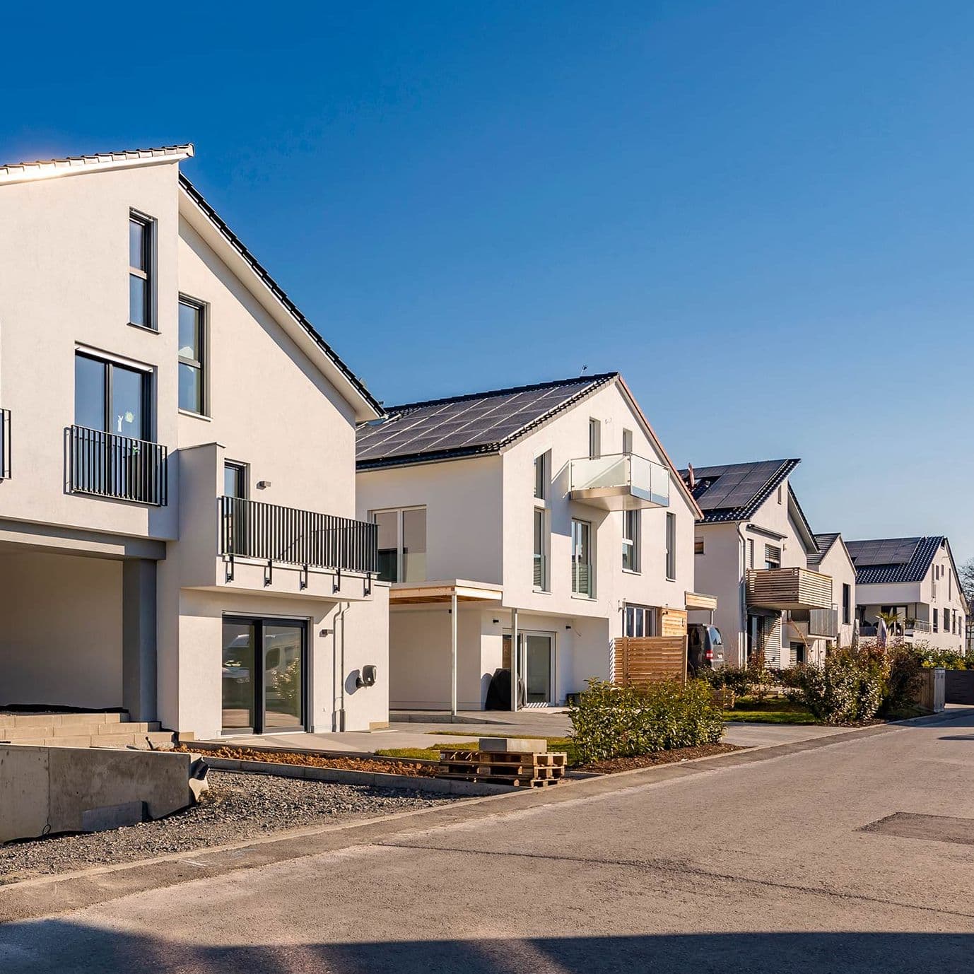 Eine Außenaufnahme eines Straßenzuges mit neugebauten Einfamilienhäusern. Ein weißes Haus ist zentral im Bild. Ähnliche Häuser reihen sich anhand einer Fluchtlinie hintereinander.