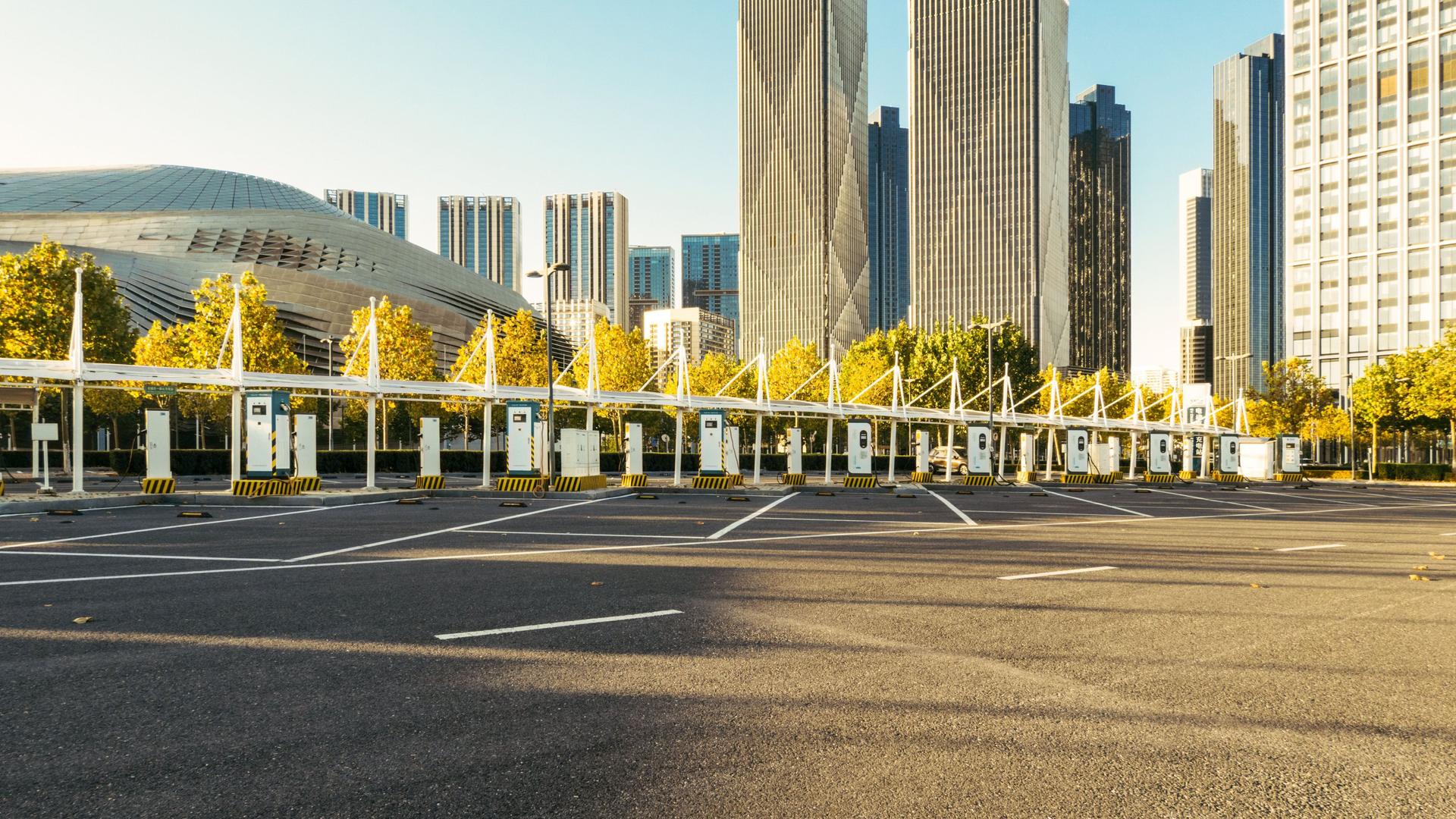Ein großer leerer Parkplatz mit Aufladestationen für Elektroautos. Im Hintergrund Hochhäuser im Sonnenschein.