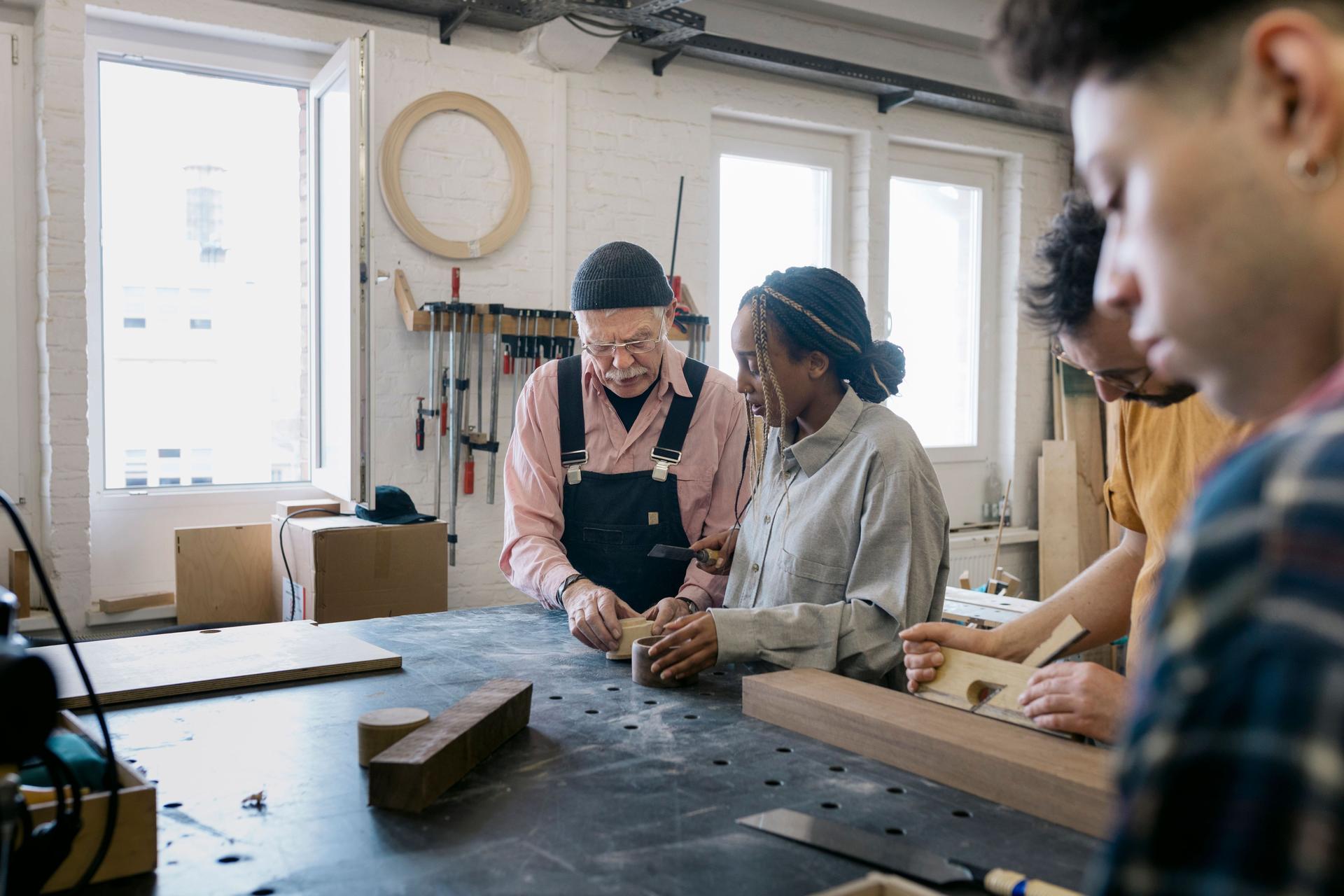 Eine junge Frau und zwei junge Männer arbeiten zusammen mit einem älteren Holzwerkstatttechniker in einer Werkstatt. Sie haben Holzblöcke vor sich.