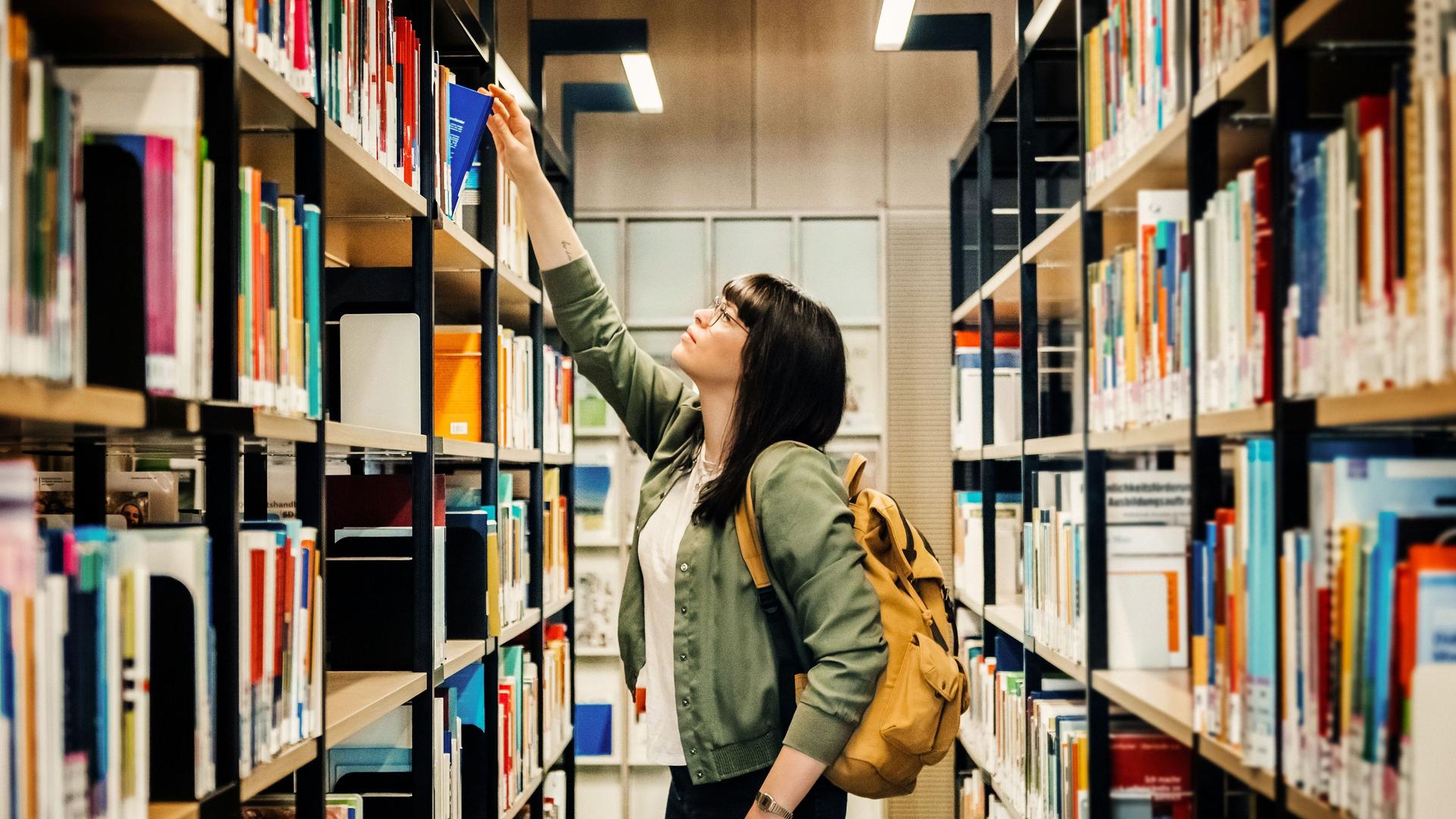 Studentin zieht in Bibliothek ein Buch aus einem Regal
