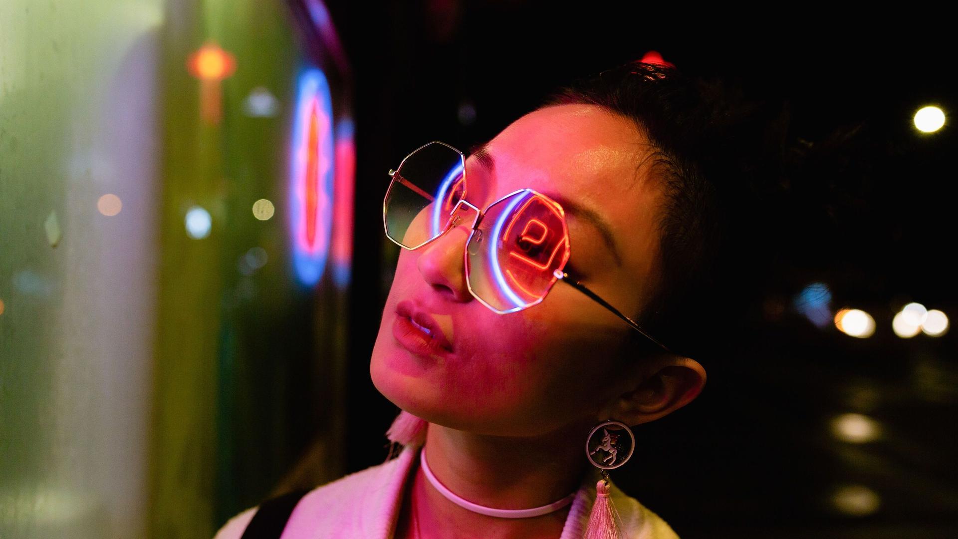 Nächtliches Porträt einer jungen Frau, die zur Seite schaut. LED Lichter reflektieren sich in ihrer großen Brille.