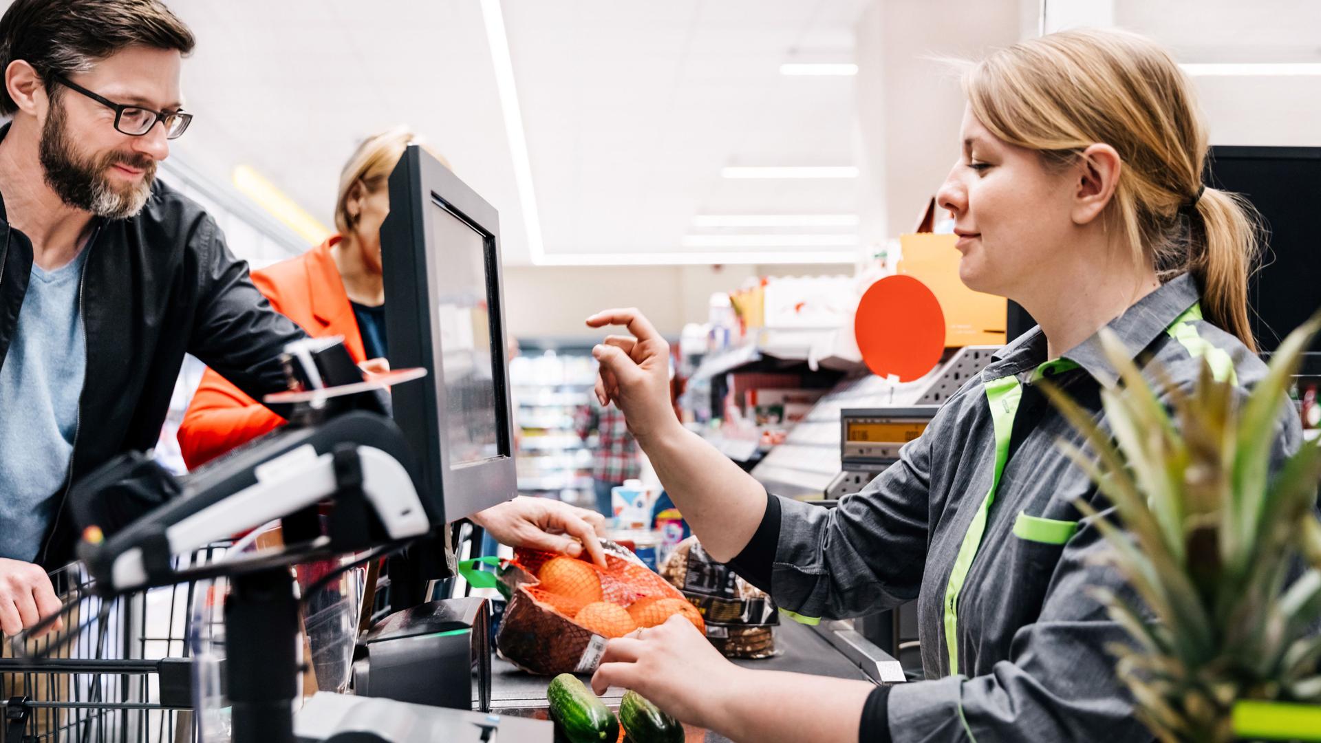 Ein Supermarktkassiererin tippt auf einen Bildschirm während zwei Gurken auf dem Scanner liegen. Ein männlicher Kunde legt derweil Waren auf das Band.