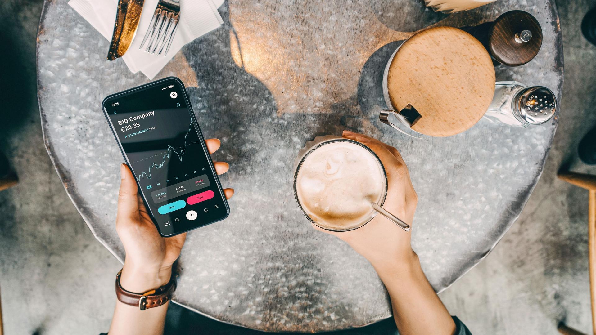 Von oben fotografierter Cafetisch. Nahaufnahme einer Person die neben einem Kaffeegetränk ein Smartphone mit Böresnkursen auf dem Bildschirm in der Hand hält.