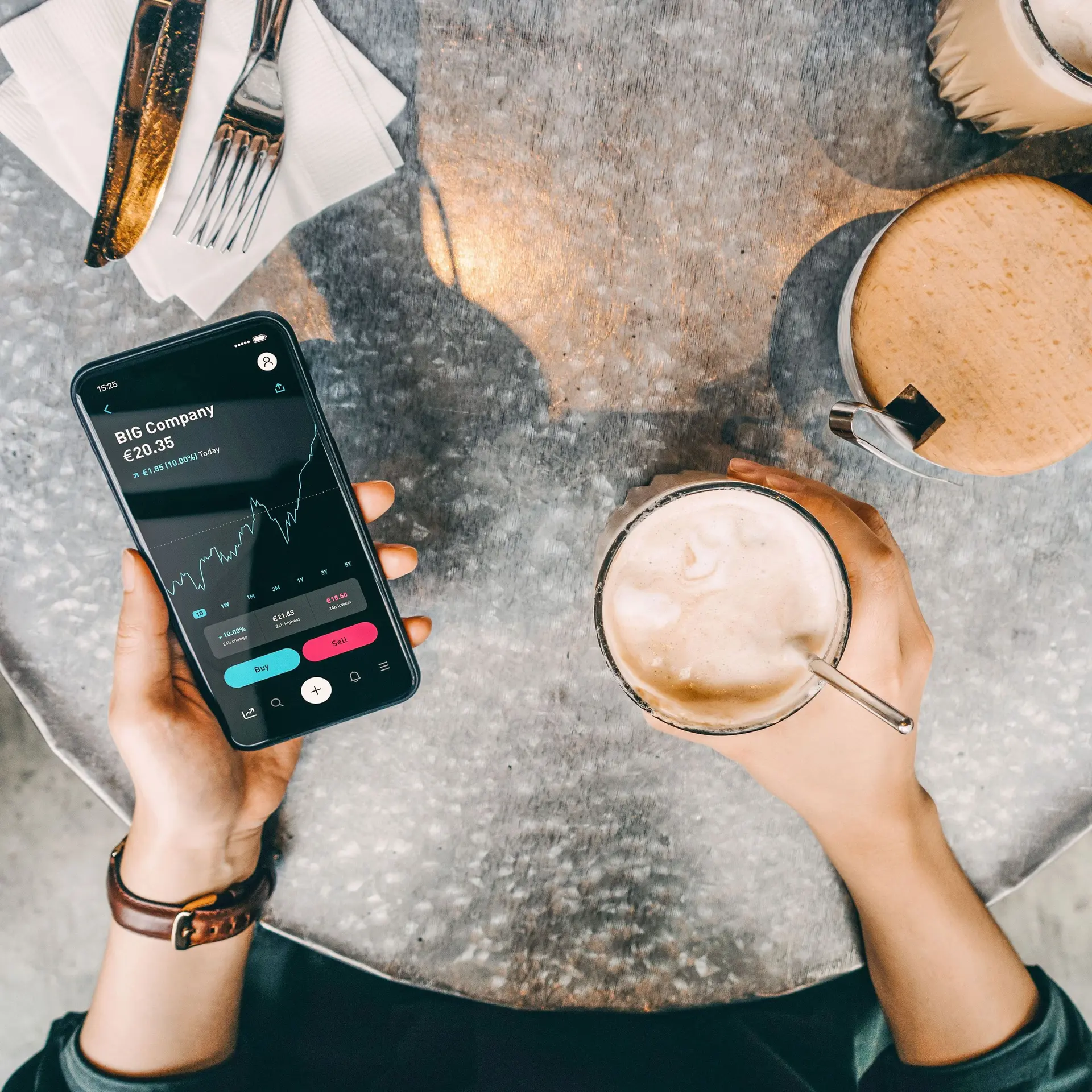 Von oben fotografierter Cafetisch. Nahaufnahme einer Person die neben einem Kaffeegetränk ein Smartphone mit Böresnkursen auf dem Bildschirm in der Hand hält.