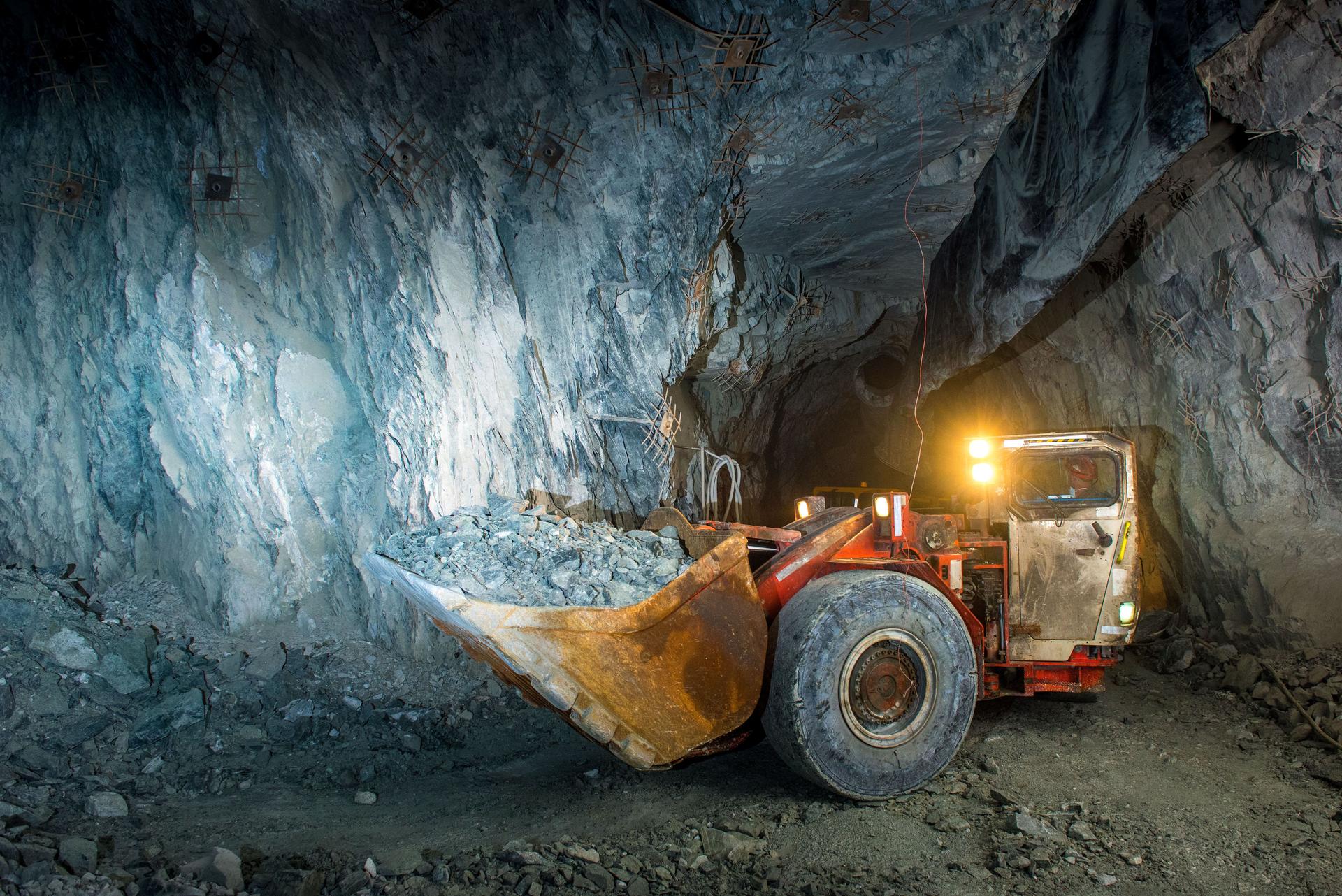 Schaufelbagger vor einer Felswand beladen mit Steinen in einem unterirdischen Tunnel.