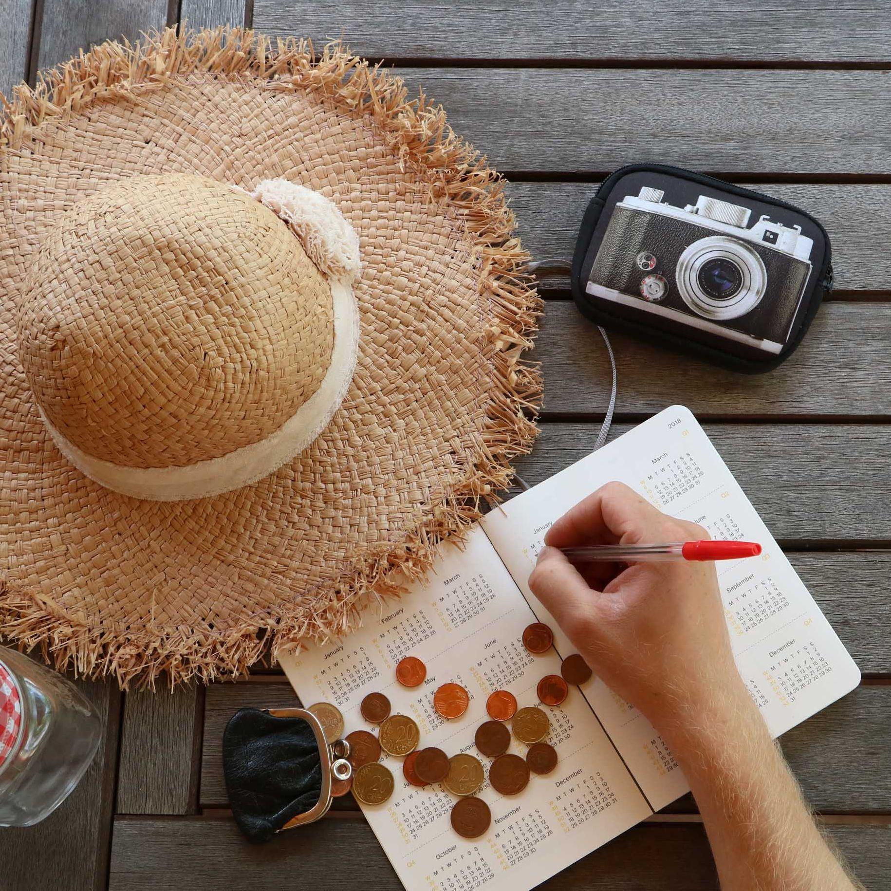 Ein Strohhut, Kleingeld, ein Trinkglas mit Deckel und Strohhalm, eine Kamera und eine Kalender liegen auf einem Tisch. Eine Hand schreibt mit einem Kugelschreiber etwas in den Kalender