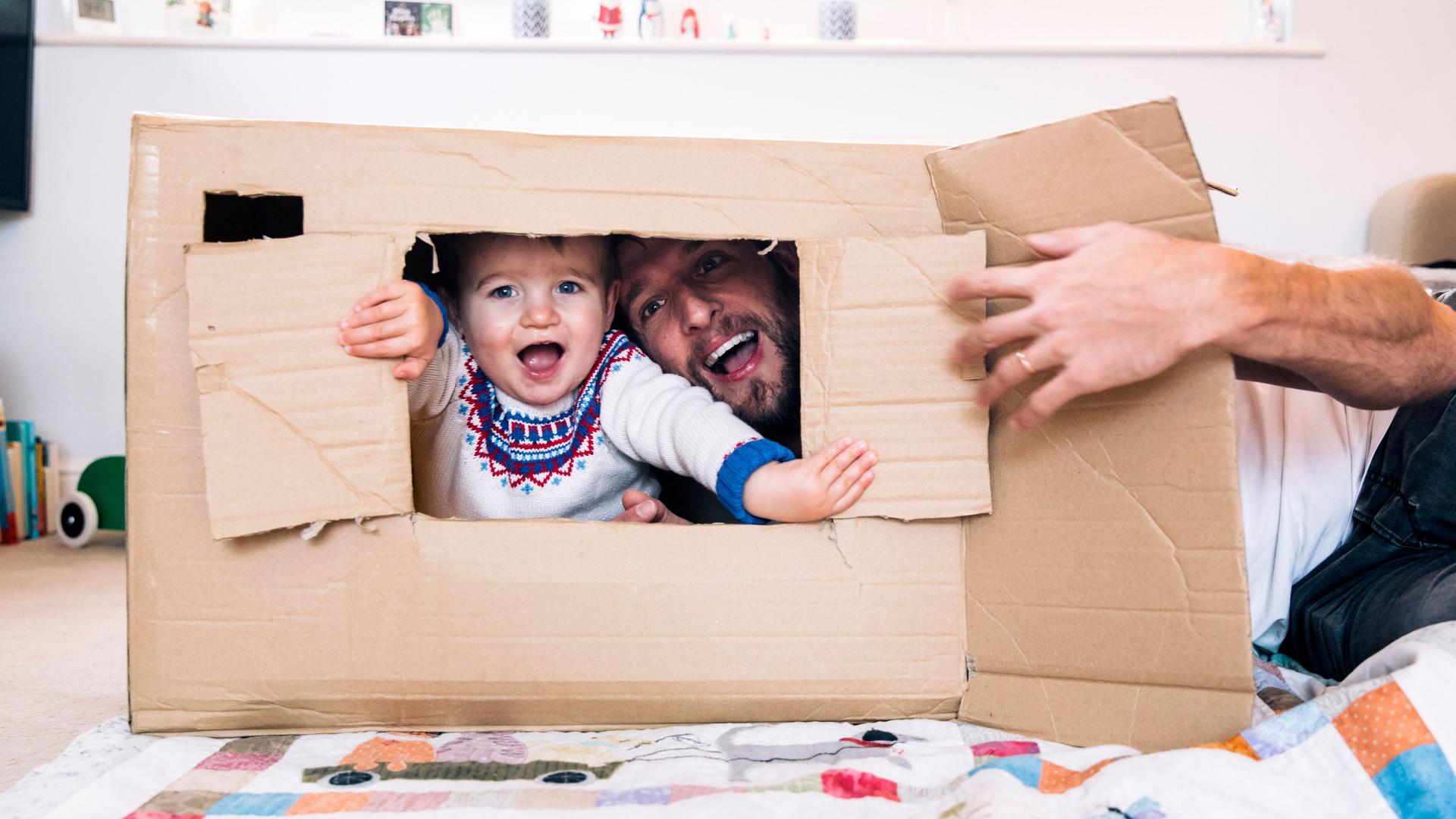 Ein Kleinkind streckt seinen Kopf aus einem Karton und spielt fröhlich mit seinem Papa.