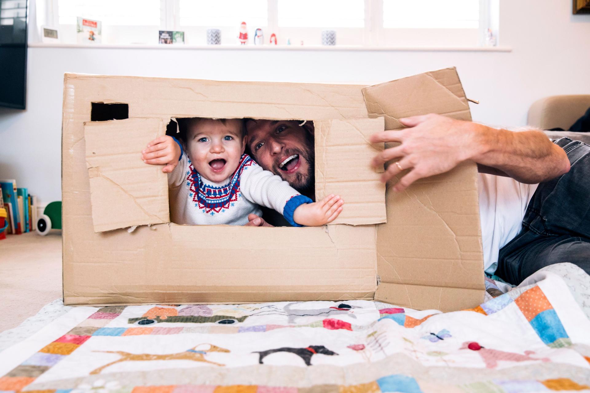 Ein Kleinkind streckt seinen Kopf aus einem Karton und spielt fröhlich mit seinem Papa.