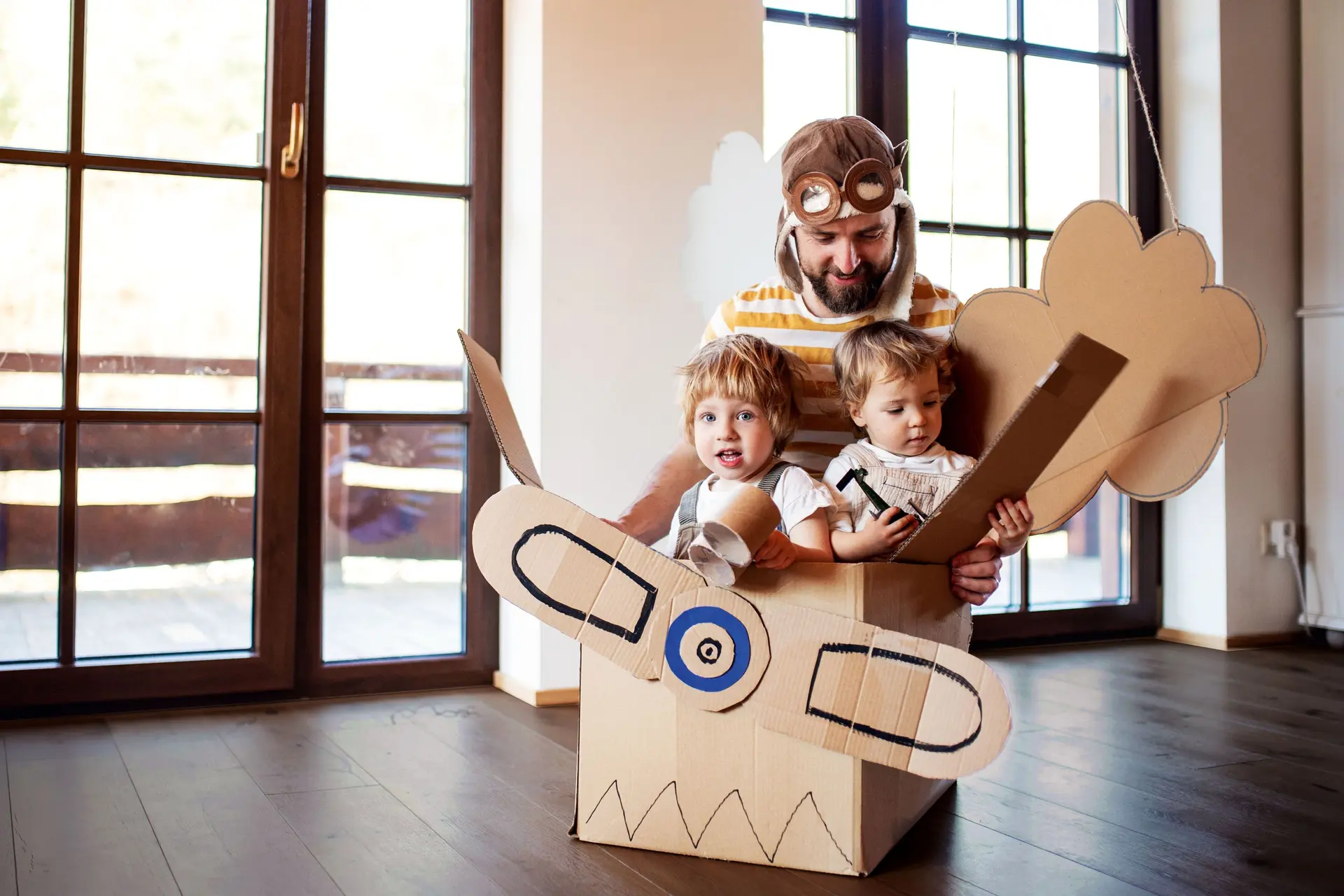 Ein Vater mit Fliegermütze spielt mit zwei Kindern in einem Flugzeug aus Pappe.